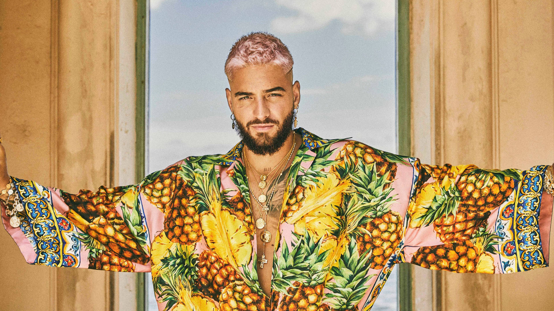 Maluma 桌布 壁紙 — 一個粉紅色頭髮和鬍鬚的男人穿著鮮豔的鳳梨印花長袍，站在窗前，雙臂伸展。