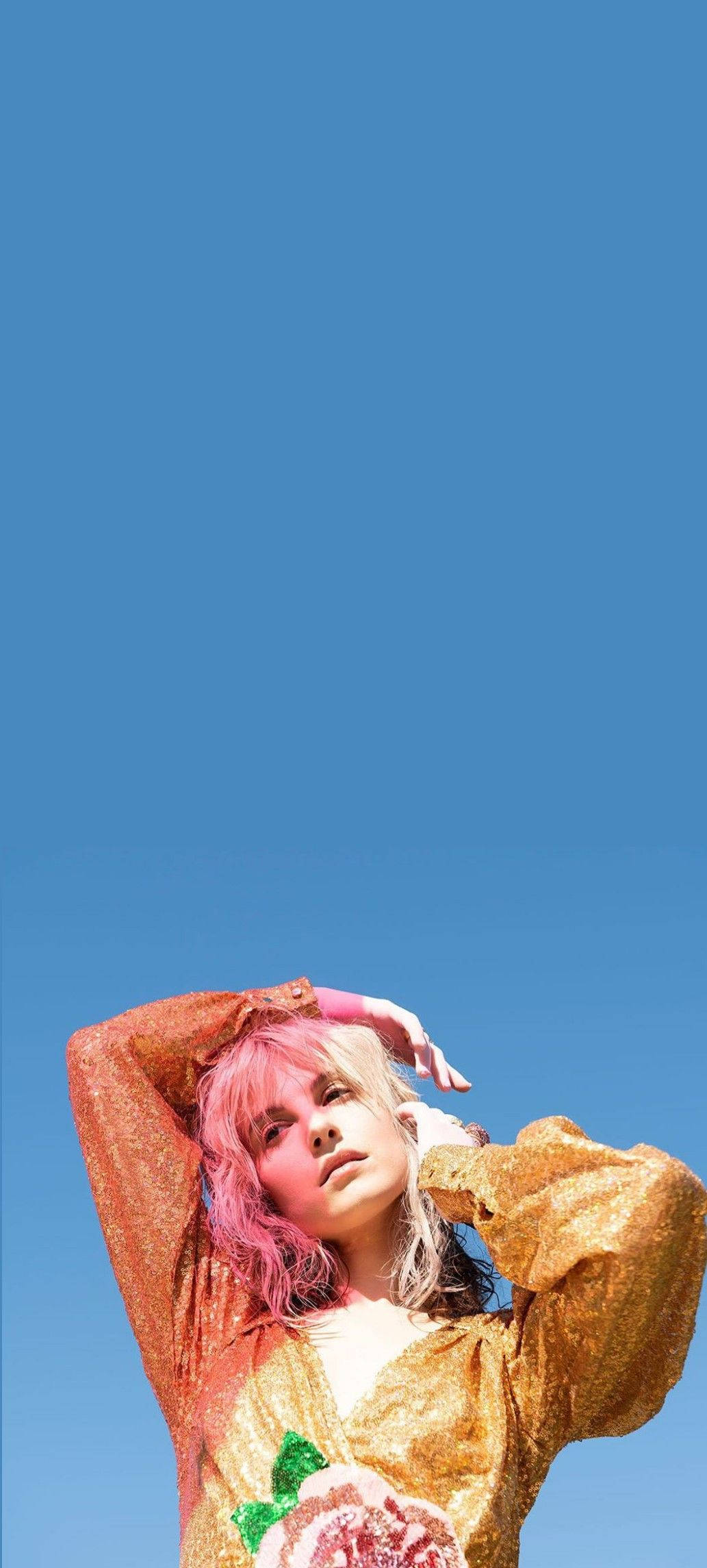 Hayley Williams 桌布 壁紙 — 一個粉紅色頭髮的人穿著閃閃發光的金色上衣，在晴朗的藍天前擺姿勢。