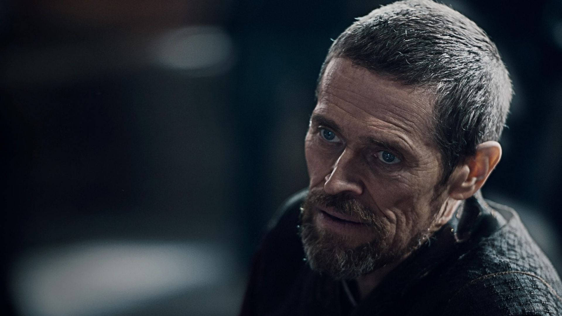 Willem Dafoe 桌布 壁紙 — 一個粗獷男人的特寫，擁有強烈的藍色眼睛和灰白的鬍鬚，在昏暗的燈光下若有所思地凝視。