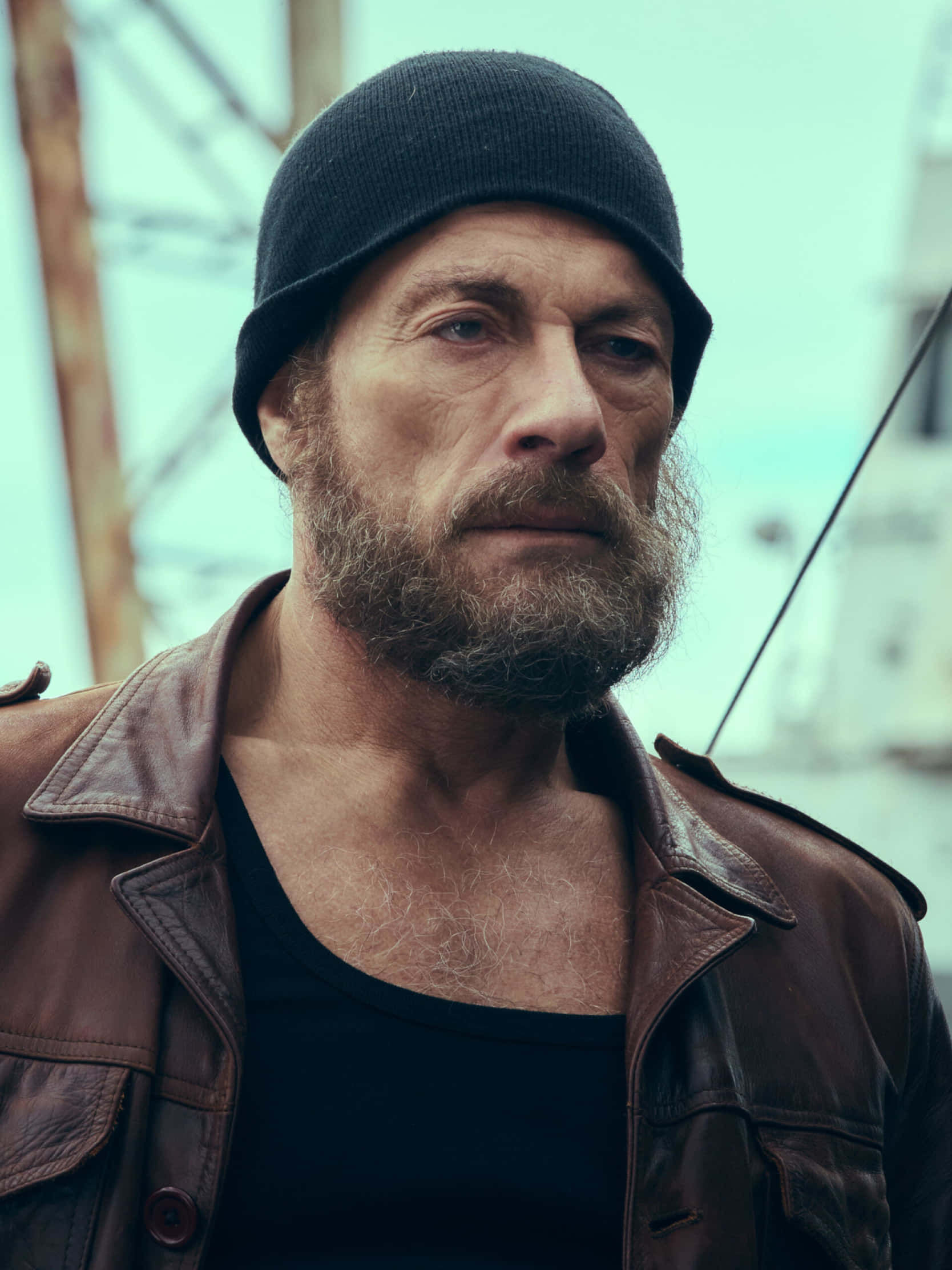 jean claude van damme 桌布 壁紙 — 一個粗獷留鬍子的男人，戴著黑色毛帽和棕色皮夾克，在模糊的工業背景下專注凝視。