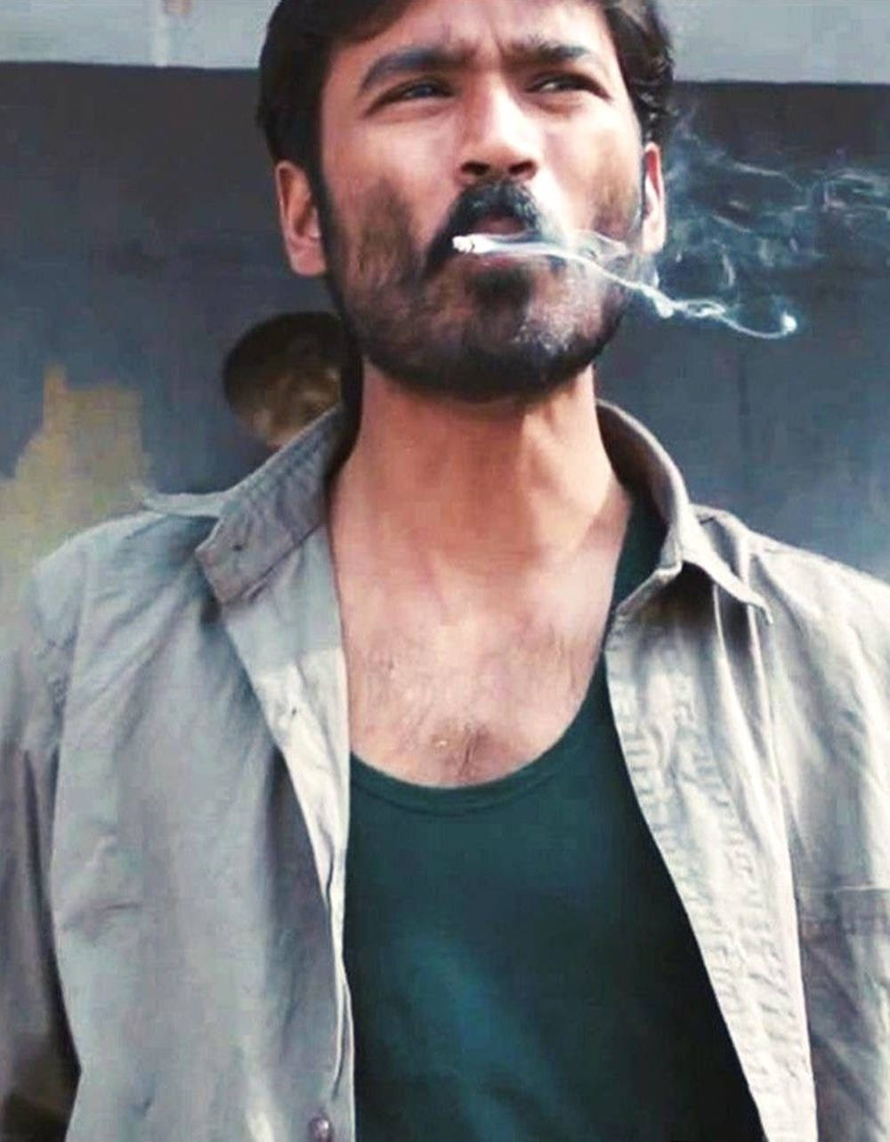 Dhanush 桌布 壁紙 — 一個粗獷留鬍子的男人抽著煙，抬頭望向模糊的城市背景。