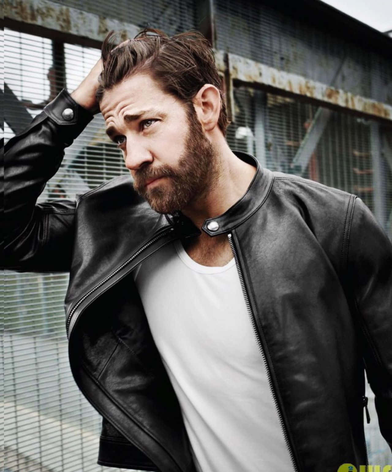 john krasinski 桌布 壁紙 — 一個粗獷留鬍的男人穿著黑色皮夾克，在生鏽的工業圍欄背景下用手撥弄頭髮。