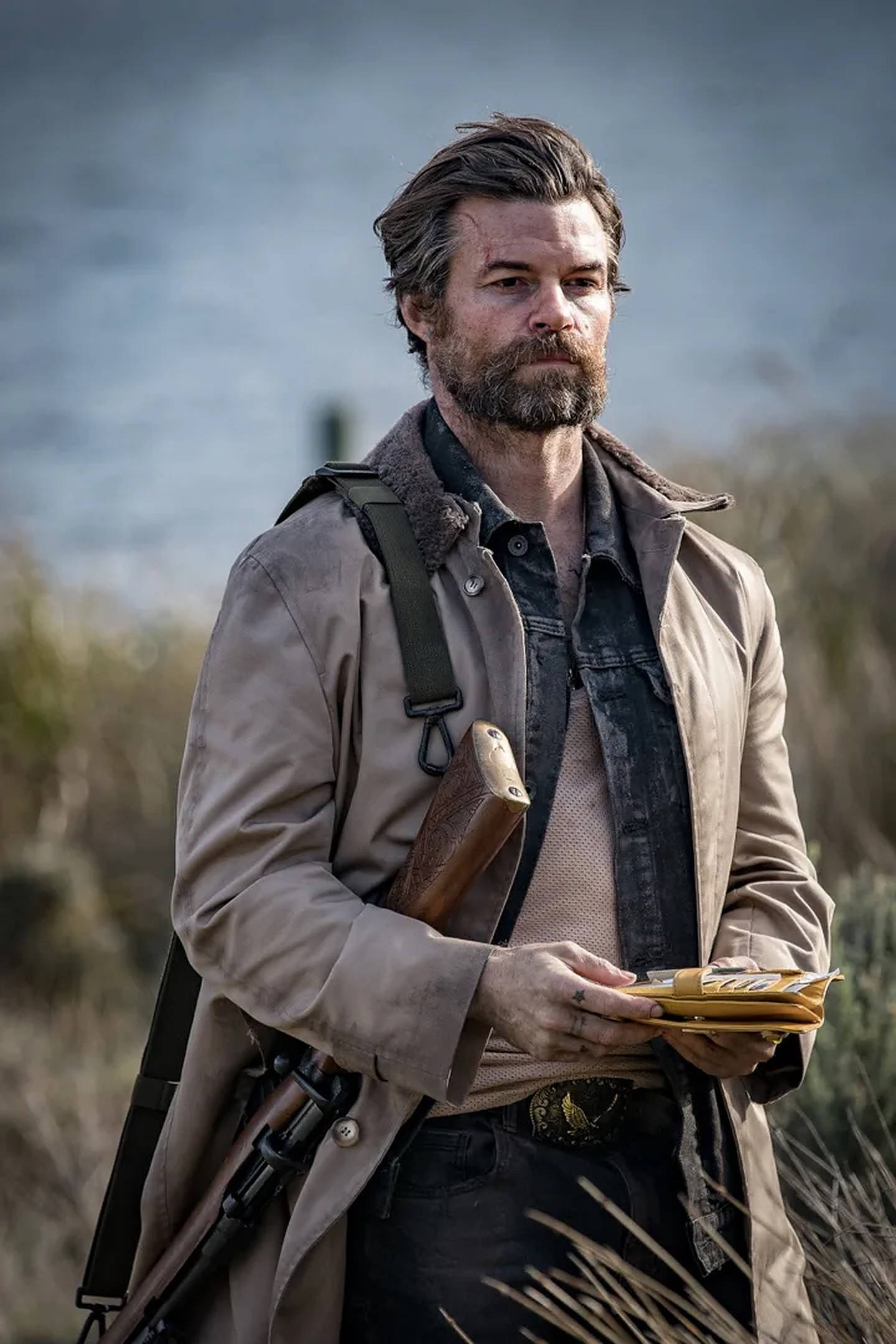 Daniel Gillies 桌布 壁紙 — 一個粗獷留鬍鬚的男人穿著米色外套，站在乾燥的草地上，手持一本皮面書，天空多雲。