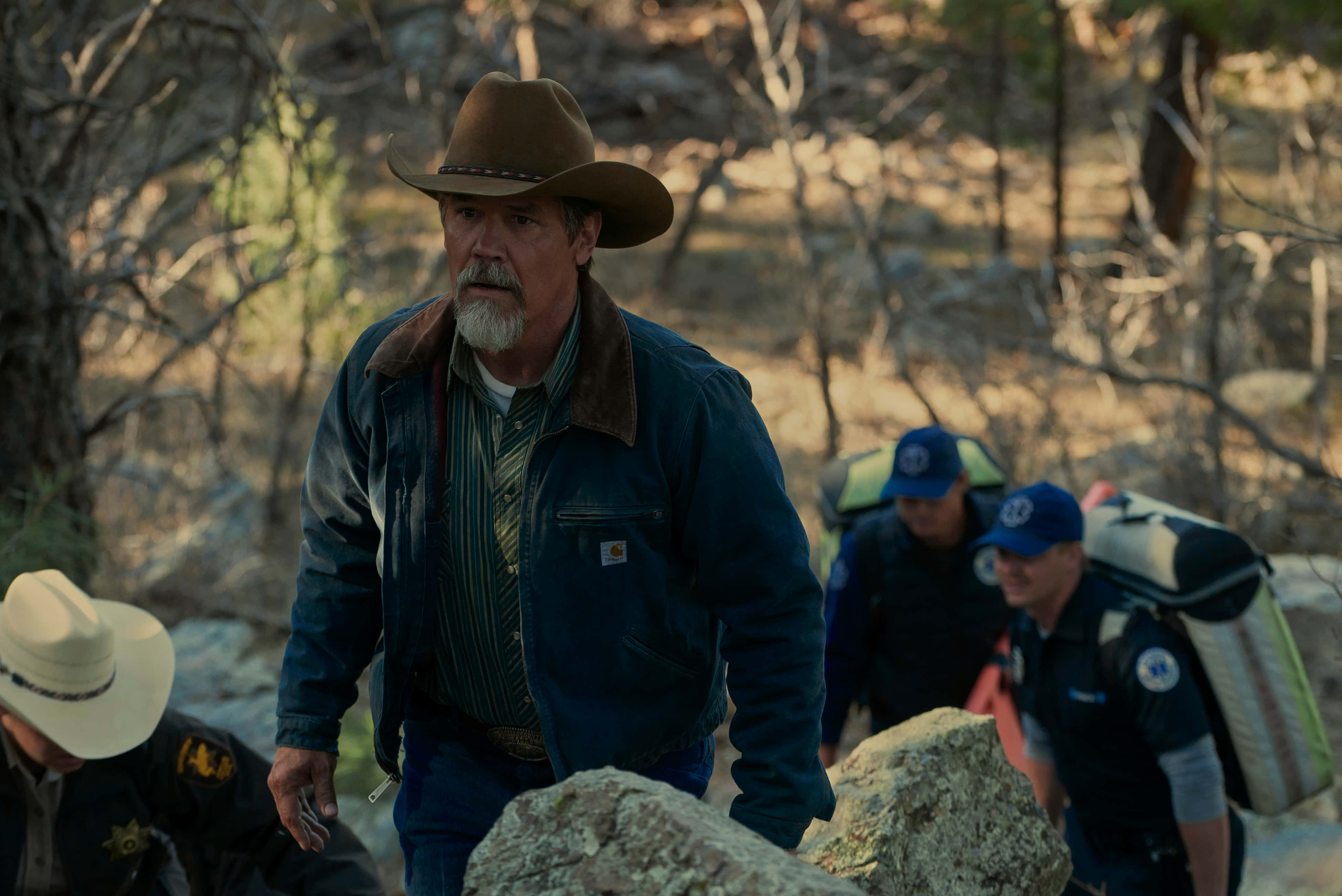 Josh Brolin 桌布 壁紙 — 一個粗獷的牛仔，留著白鬍子，戴著棕色帽子，站在陽光普照的森林中，身後有帶著裝備的救援人員跟隨。