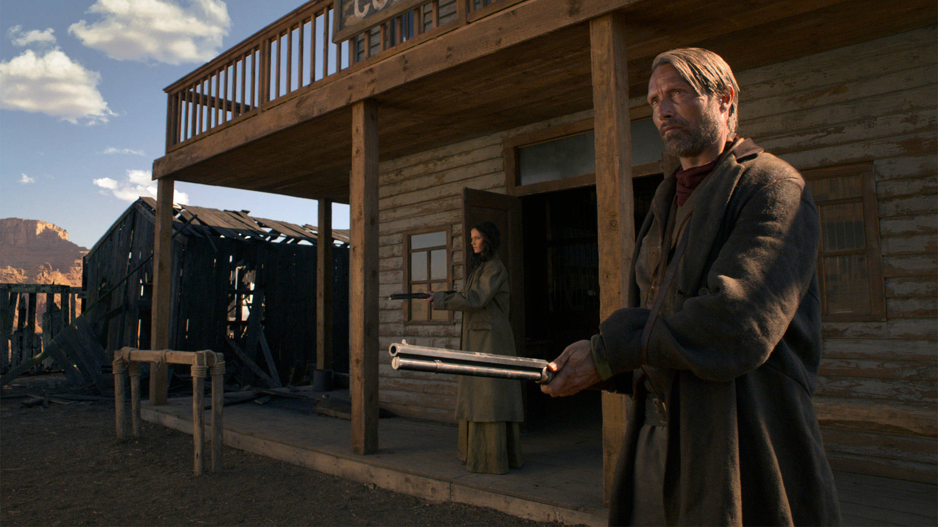 Mads Mikkelsen 桌布 壁紙 — 一個粗獷的牛仔站在塵土飛揚的沙龍門廊上，手持步槍，而一名女子在門口用手槍瞄準，頭頂是明亮的藍天。