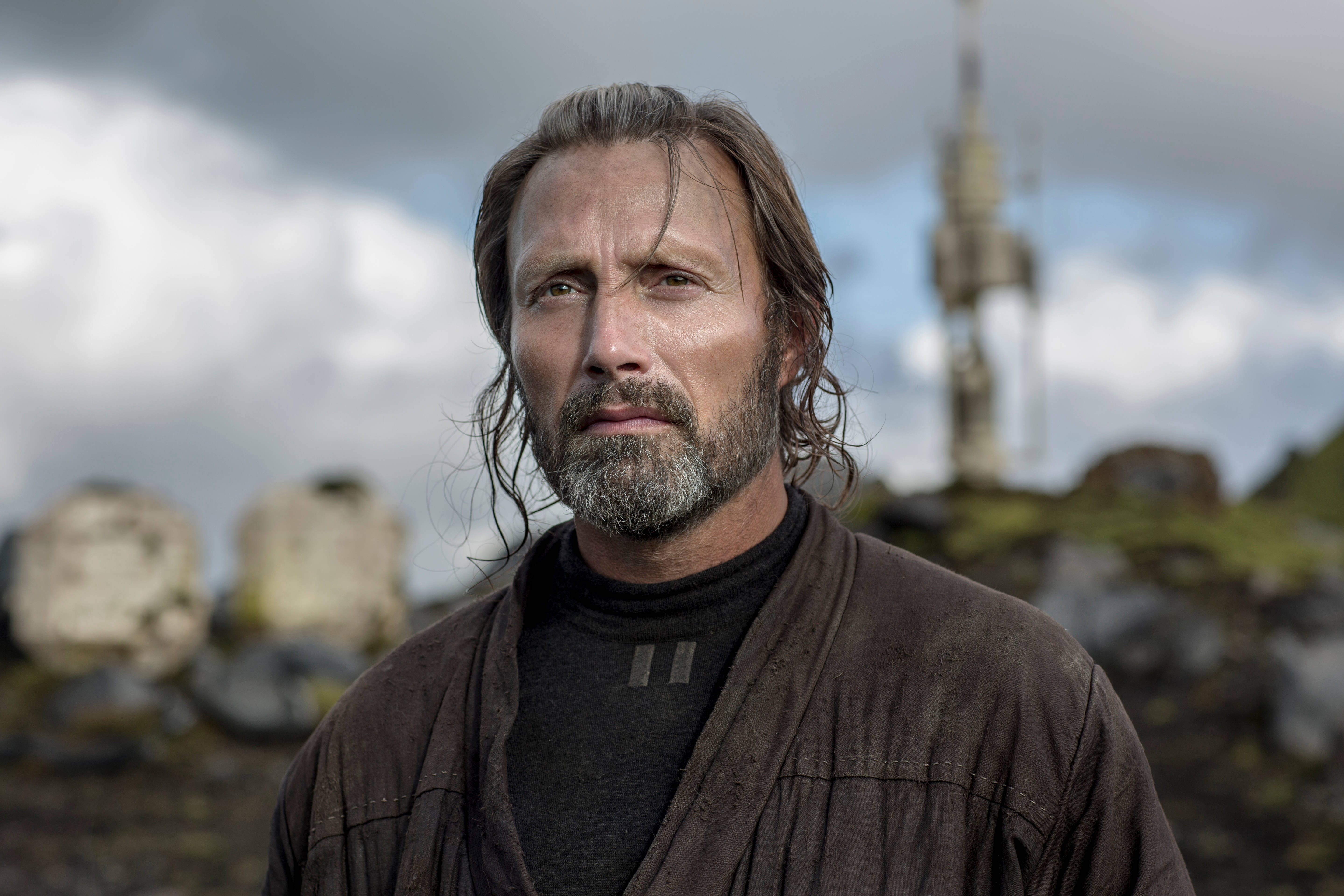 Mads Mikkelsen 桌布 壁紙 — 一個粗獷的男人，留著風霜鬍鬚，眼神銳利，站在戶外，背景是陰天和遠處的塔樓。