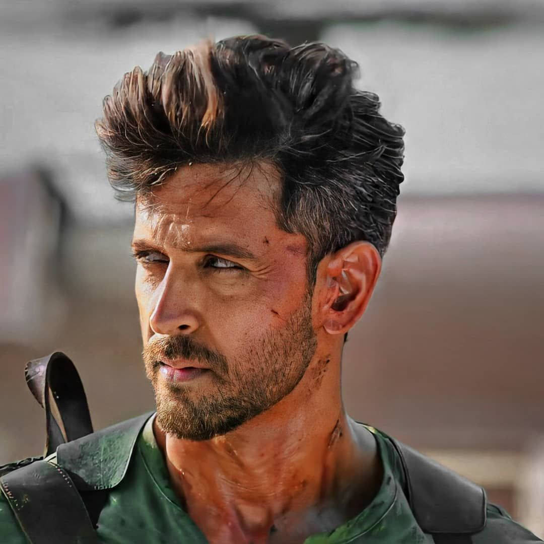 Indian 桌布 壁紙 — 一個粗獷的男人，頭髮凌亂、深色，留著鬍鬚，穿著一件綠色軍裝風格襯衫，肩上掛著一條帶子，專注地向前凝視。