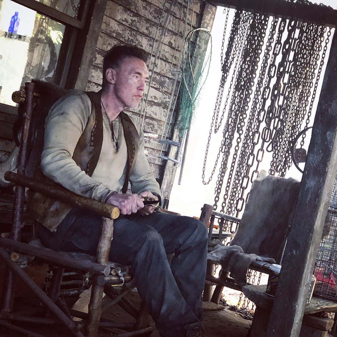 Kevin Durand 桌布 壁紙 — 一個粗獷的男人坐在一間破舊棚屋內一張風化的木椅上，棚屋內掛著鏈條和漁網。