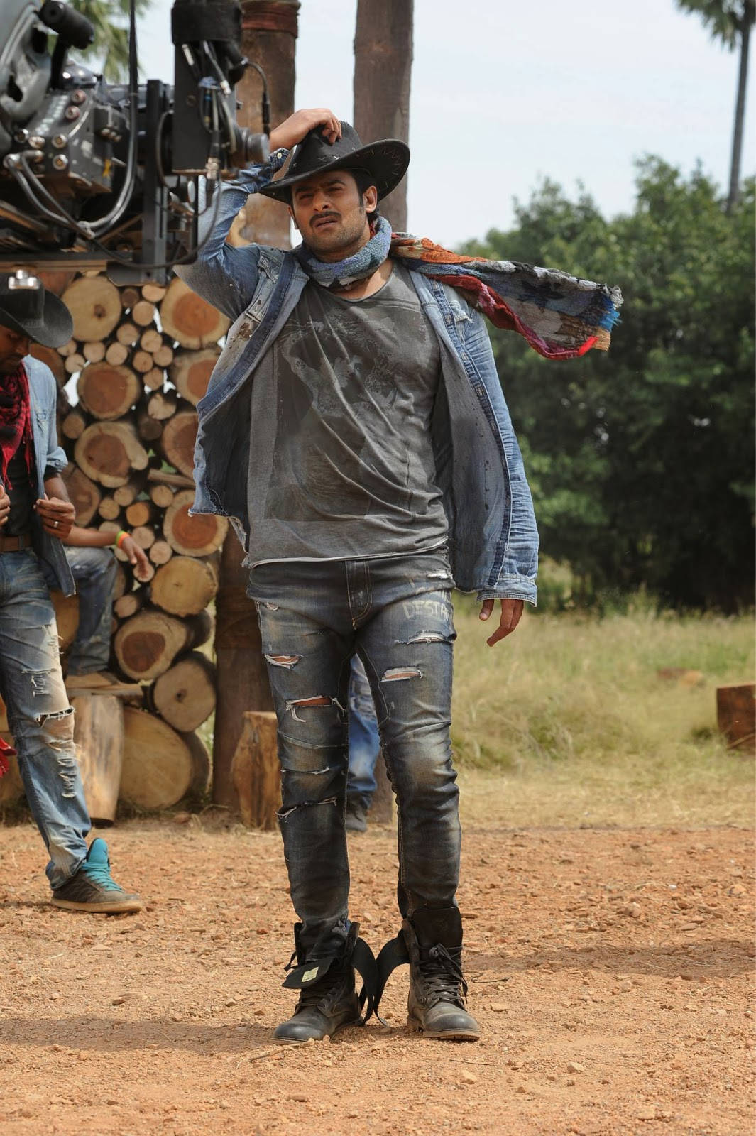 Prabhas Mirchi 桌布 壁紙 — 一個粗獷的男人戴著牛仔帽和破洞牛仔褲，自信地站在一條土路上，旁邊是一堆堆疊的木柴，天空明亮。