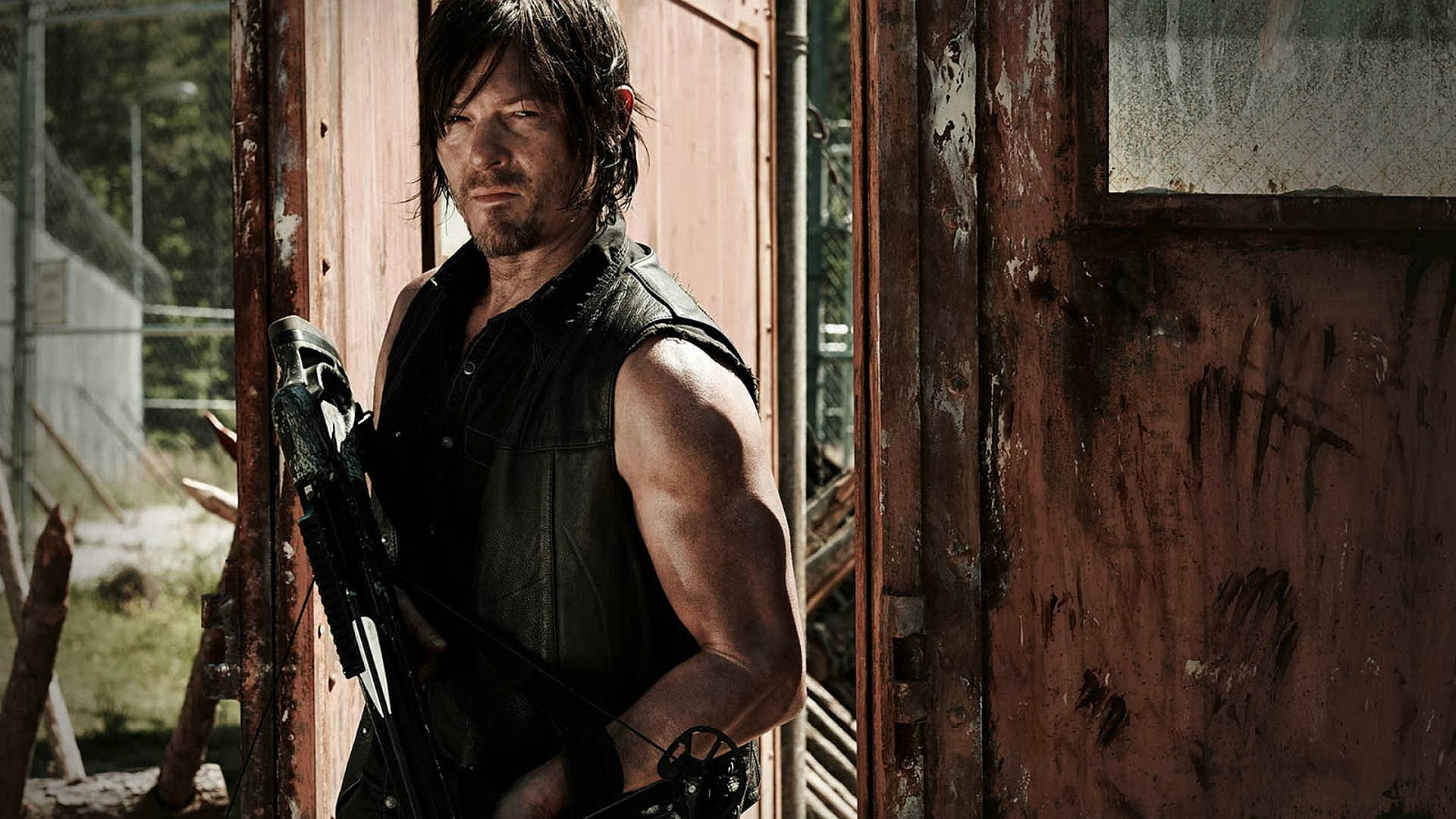 Walking Dead Daryl 桌布 壁紙 — 一個粗獷的男人拿著弓站在風化的木門前，陽光在他堅定的臉上投下陰影。
