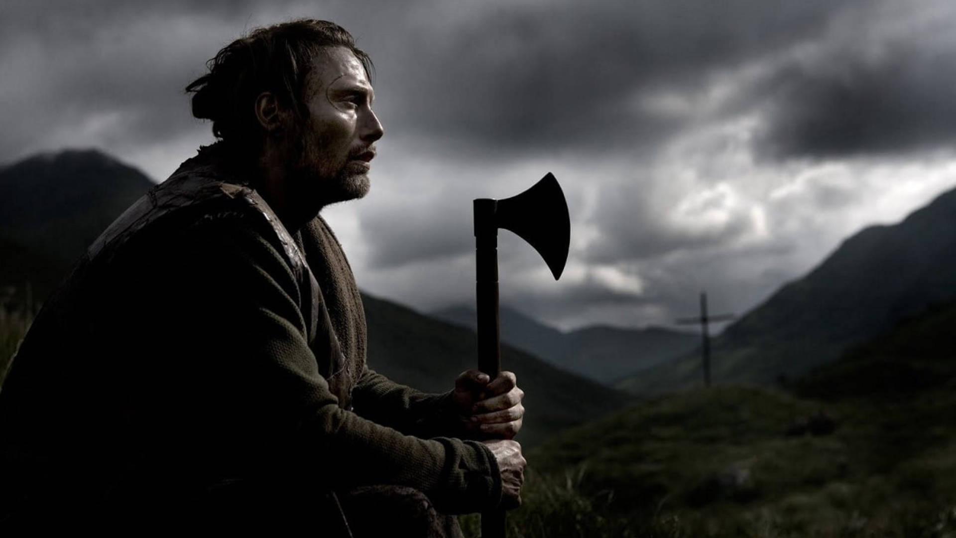 Mads Mikkelsen 桌布 壁紙 — 一個粗獷的男人拿著斧頭，在黑暗的雲層下的暴風雨山景中形成剪影。
