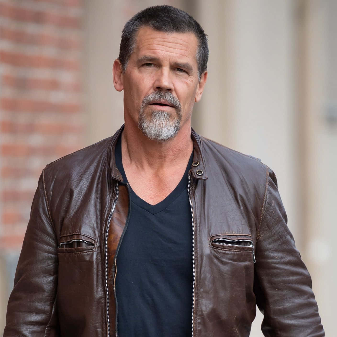 Josh Brolin 桌布 壁紙 — 一個粗獷的男人留著灰白鬍鬚，身穿棕色皮夾克內搭黑色T恤，背景是磚牆。