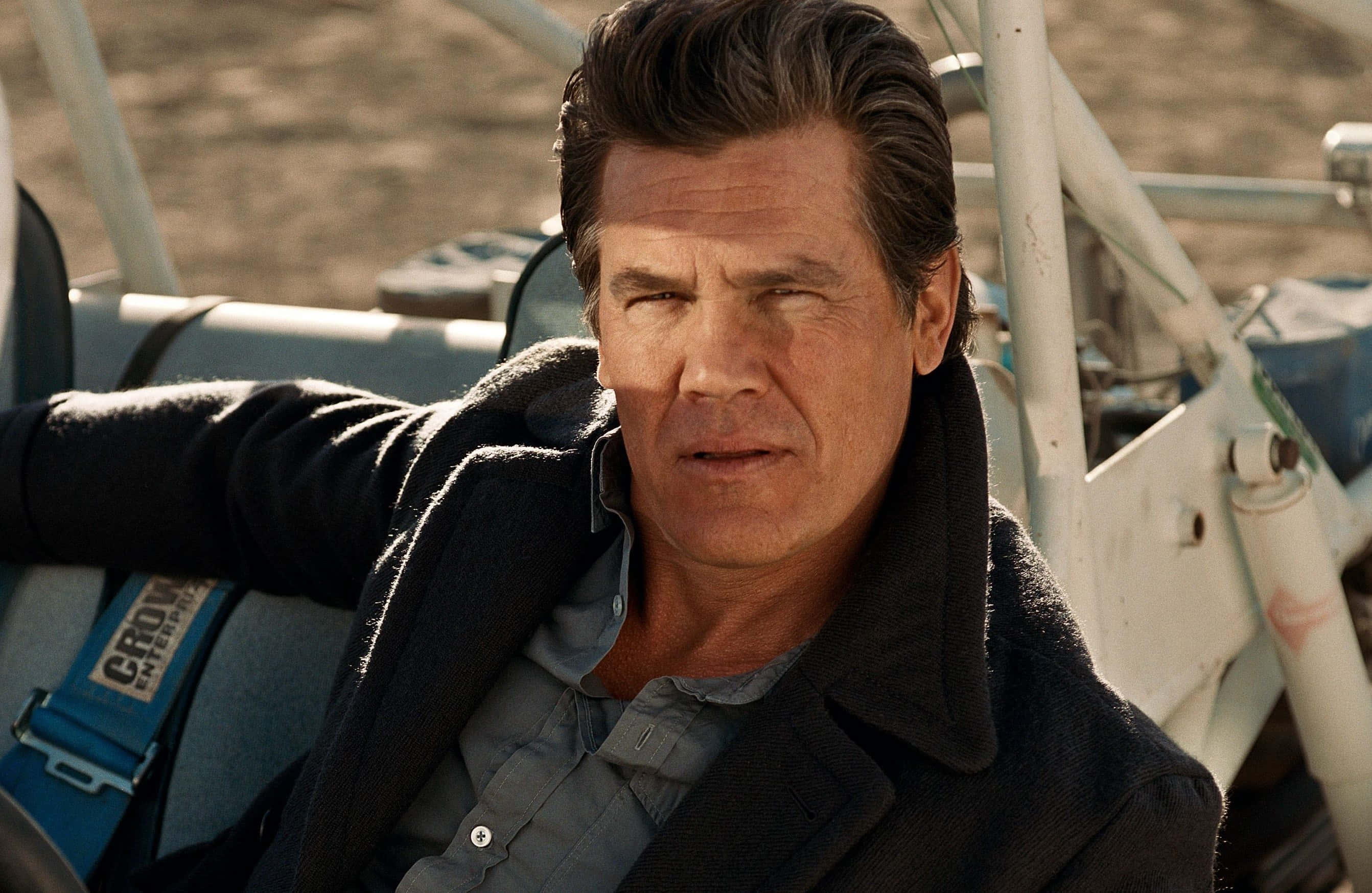 Josh Brolin 桌布 壁紙 — 一個粗獷的男人穿著深色外套，在明亮的沙漠陽光下靠著沙灘車，身後是塵土飛揚的地形。