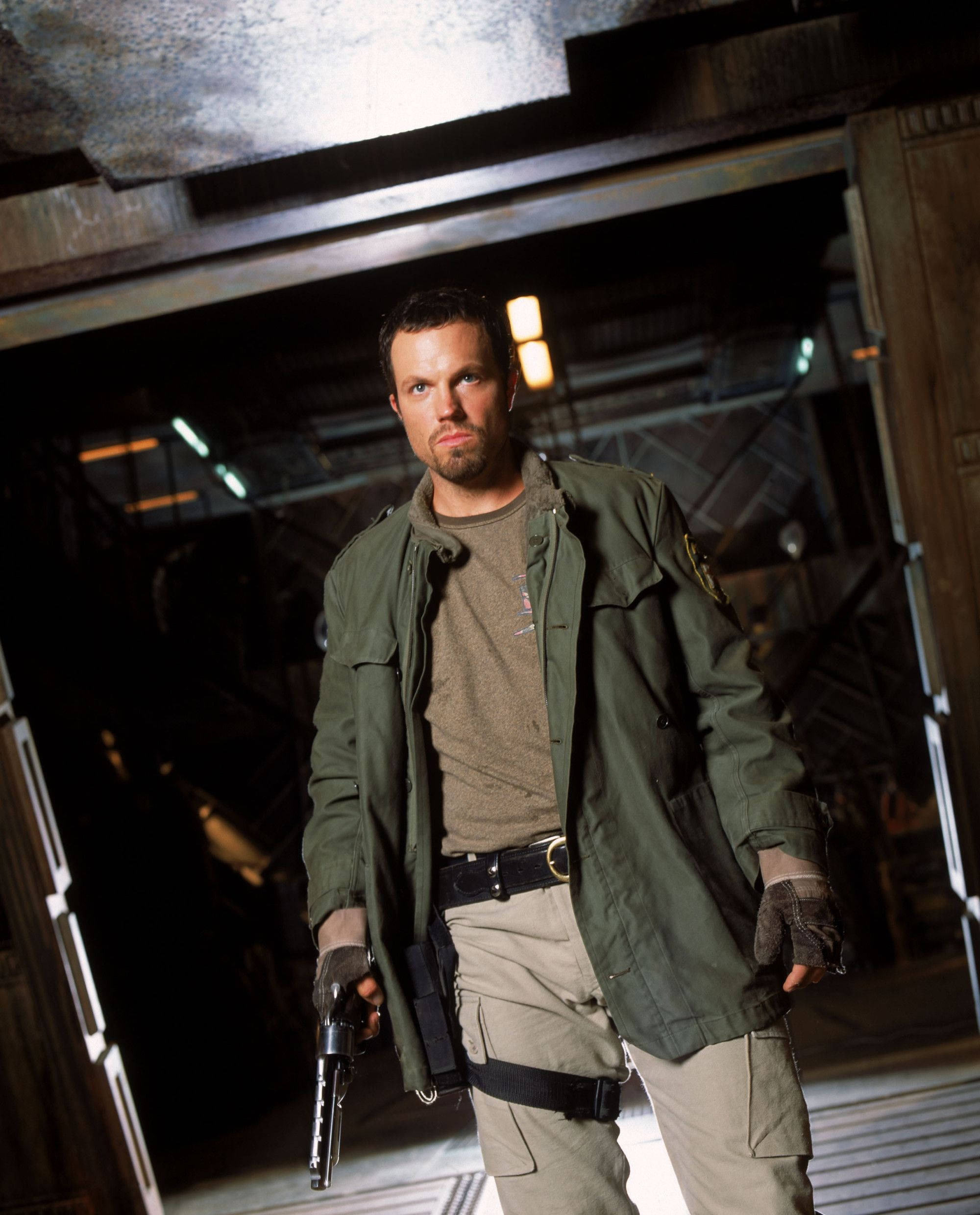 adam baldwin 桌布 壁紙 — 一個粗獷的男人穿著綠色軍用外套，在燈光昏暗的工業機庫中手持手槍，上方有燈光。