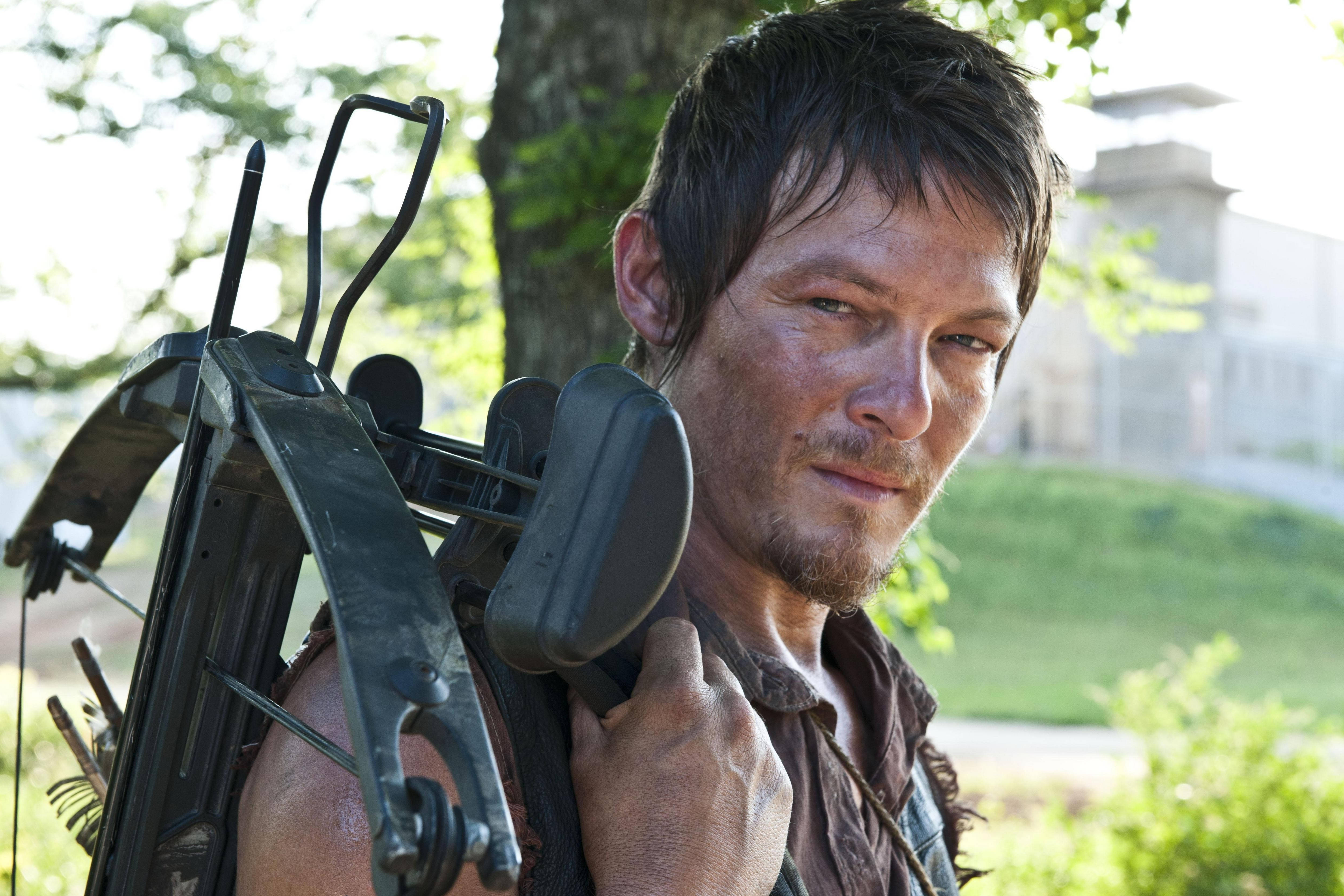 Walking Dead Daryl 桌布 壁紙 — 一個粗獷的男人肩上掛著十字弓，站在陽光明媚的戶外環境中，背景有樹木和建築物。