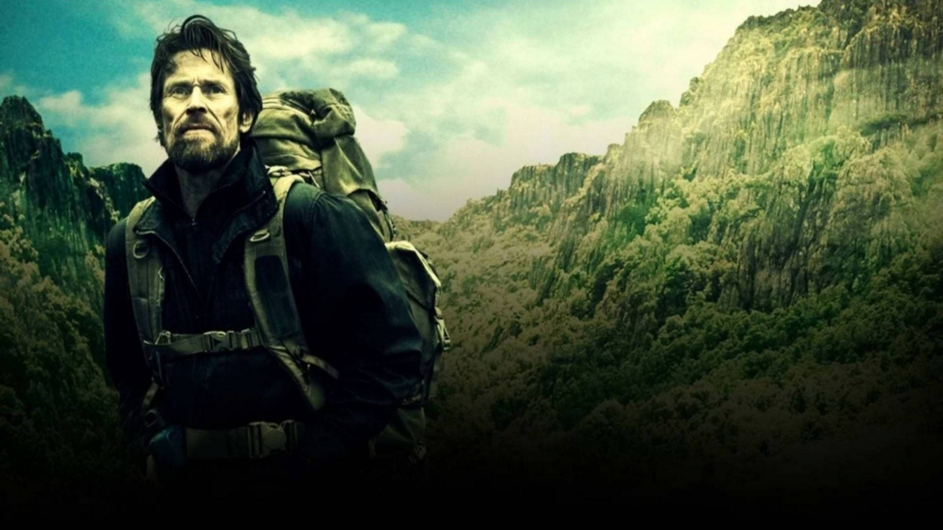 Willem Dafoe 桌布 壁紙 — 一個粗獷的鬍鬚男背著一個大背包，在陰天背景下，以高聳的綠色山脈為背景，莊嚴地站立。