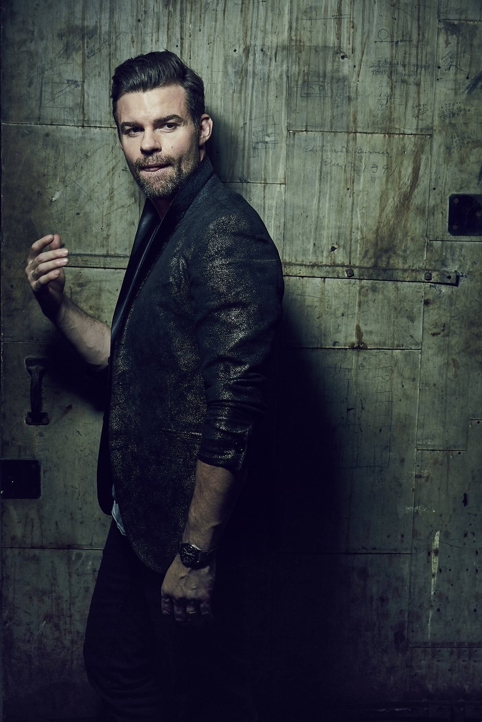 Daniel Gillies 桌布 壁紙 — 一個粗獷英俊、留鬍鬚的男人靠在一扇飽經風霜的金屬門上，環境陰暗、燈光昏暗。