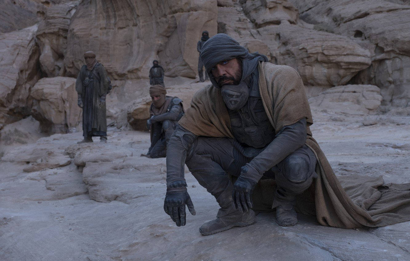 Dune Movie 桌布 壁紙 — 一個粗糙的沙漠場景，一名戴著兜帽的戰士蹲在岩石峽谷中，而其他人站在背景中。