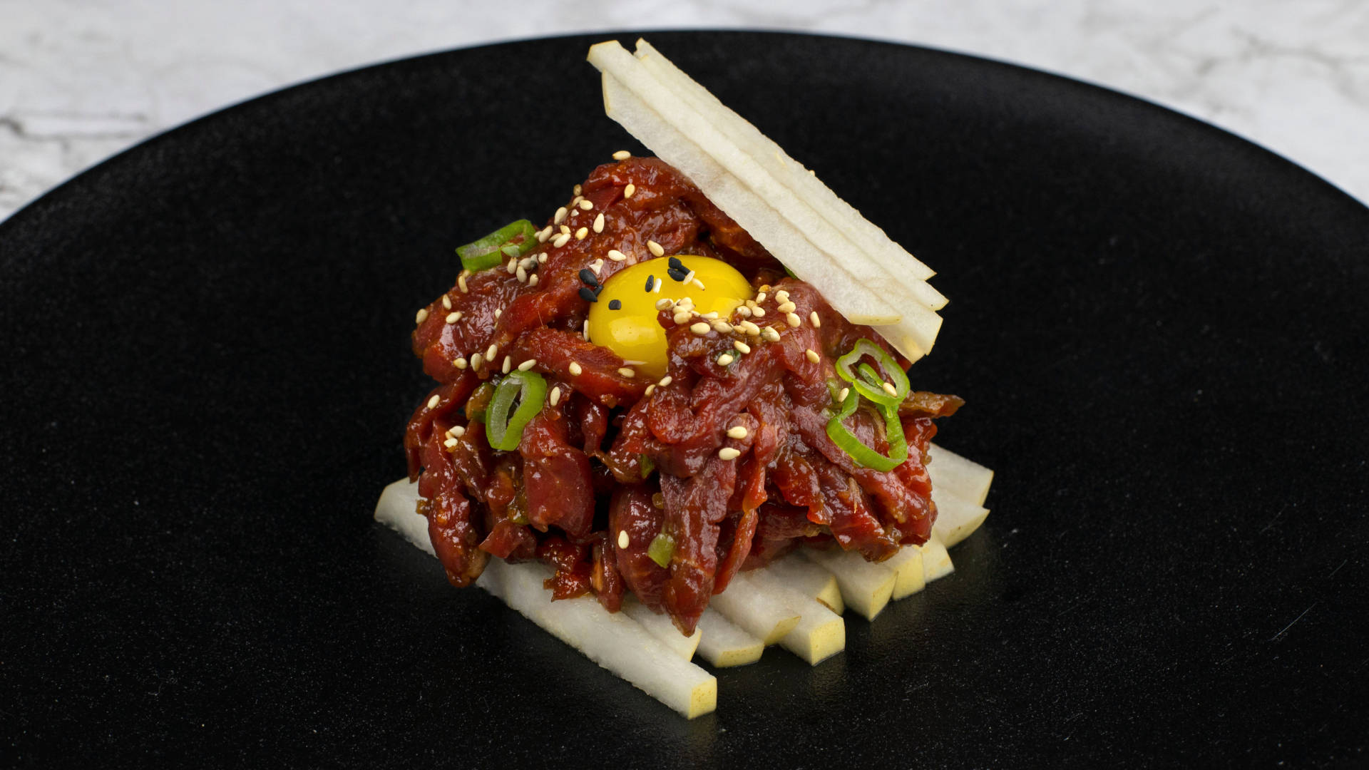 meat 桌布 壁紙 — 一個精心擺盤的菜餚，以黑盤盛載辛辣醃製牛肉，上面放有生蛋黃和芝麻。