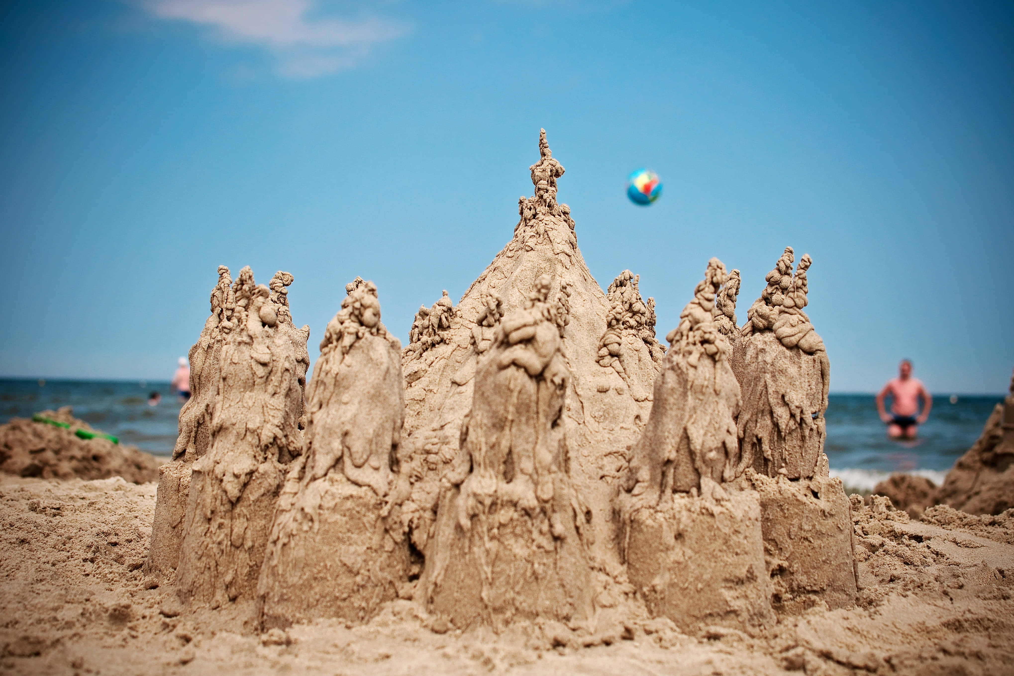 Sandcastles 桌布 壁紙 — 一個精細的沙堡，帶有塔樓和人物，矗立在陽光明媚的海灘上，一個色彩繽紛的沙灘球在空中漂浮。
