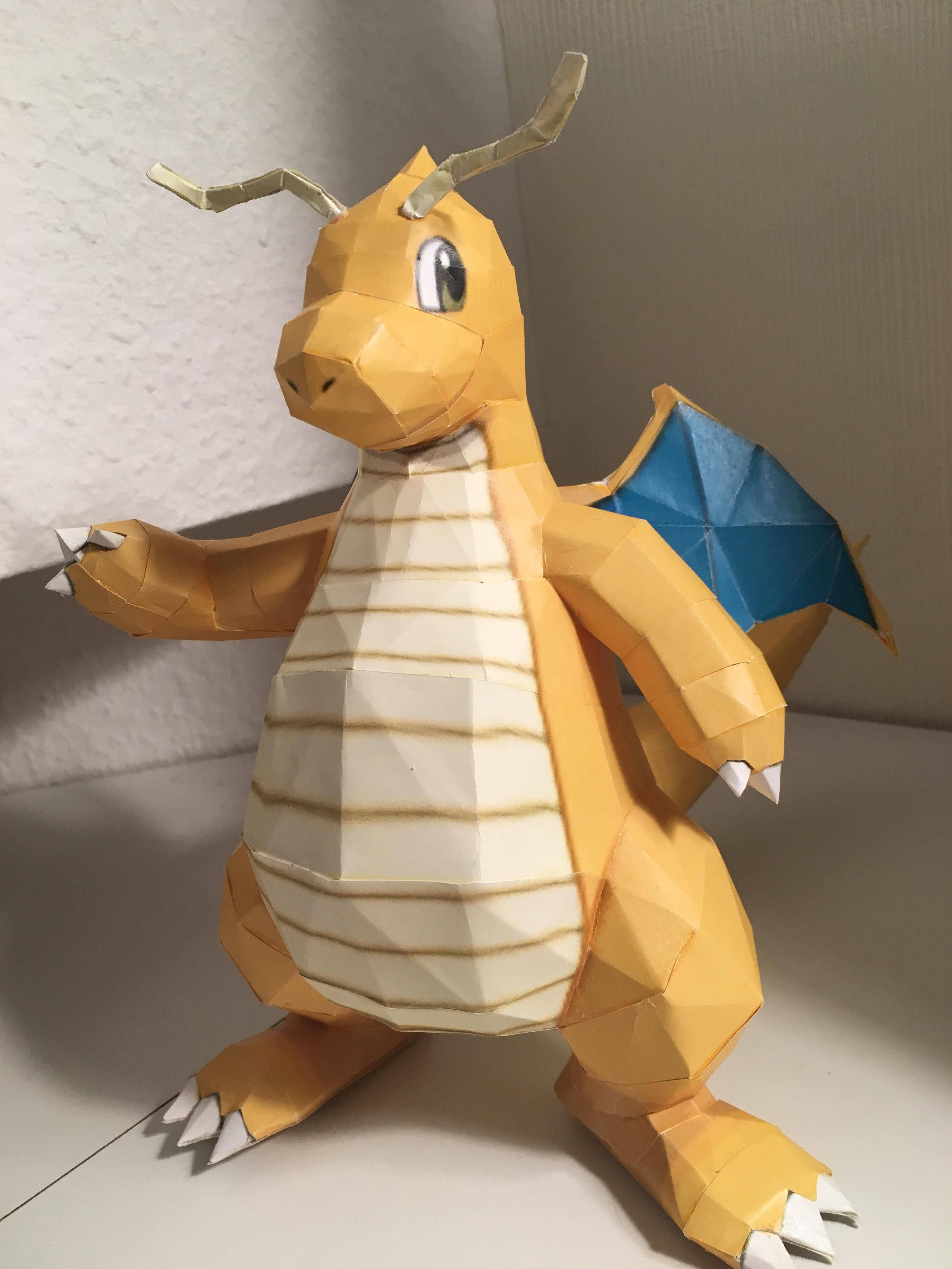 Dragonite 桌布 壁紙 — 一個精細的紙藝模型，寶可夢噴火龍擺出姿勢，擁有藍色翅膀和白色爪子，背景是素牆。