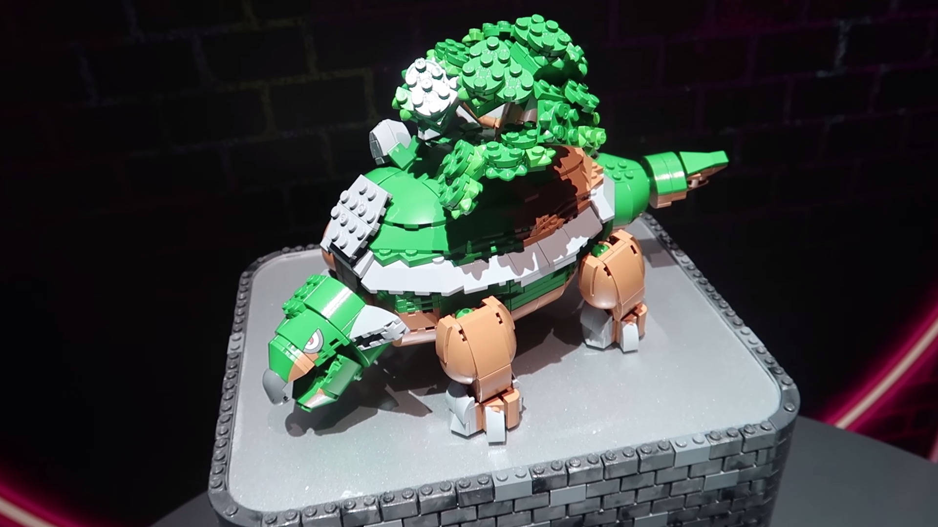 寶可夢 桌布 壁紙 — 一個精細的綠色和棕褐色LEGO恐龍模型站在灰色磚砌底座上，背景是帶有霓虹燈照明的深色磚牆。