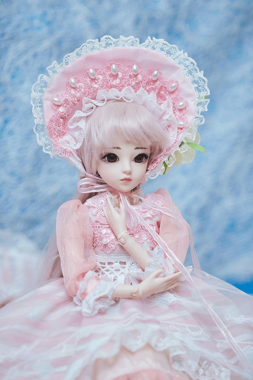 Doll 桌布 壁紙 — 一個精緻的洋娃娃，身穿粉紅色蕾絲長裙和飾有珍珠的帽子，在柔和的藍色紋理背景前優雅地擺姿勢。