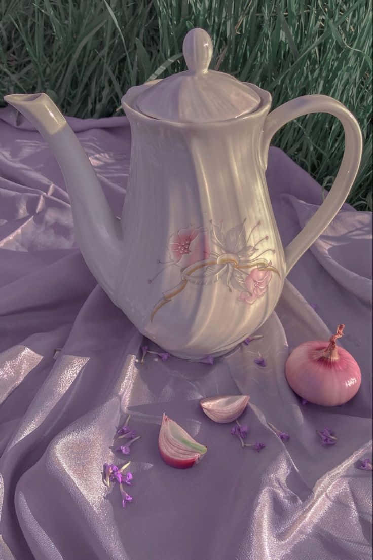 Violet Aesthetic 桌布 壁紙 — 一個精緻的白色茶壺帶有花卉貼花，放在垂墜的薰衣草布料上，旁邊有一個完整和切片的粉紅色洋蔥和散落的紫色花朵。
