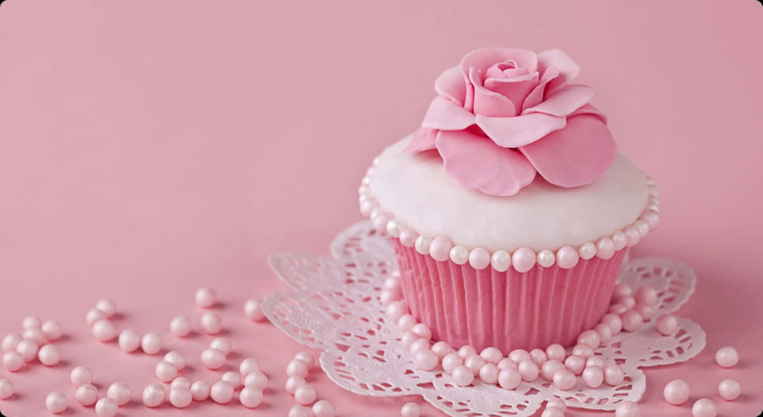 Cute Cupcake 桌布 壁紙 — 一個精緻的粉紅色杯子蛋糕，上面有糖玫瑰和珍珠糖粒，放在蕾絲墊上，背景是柔和的粉紅色。