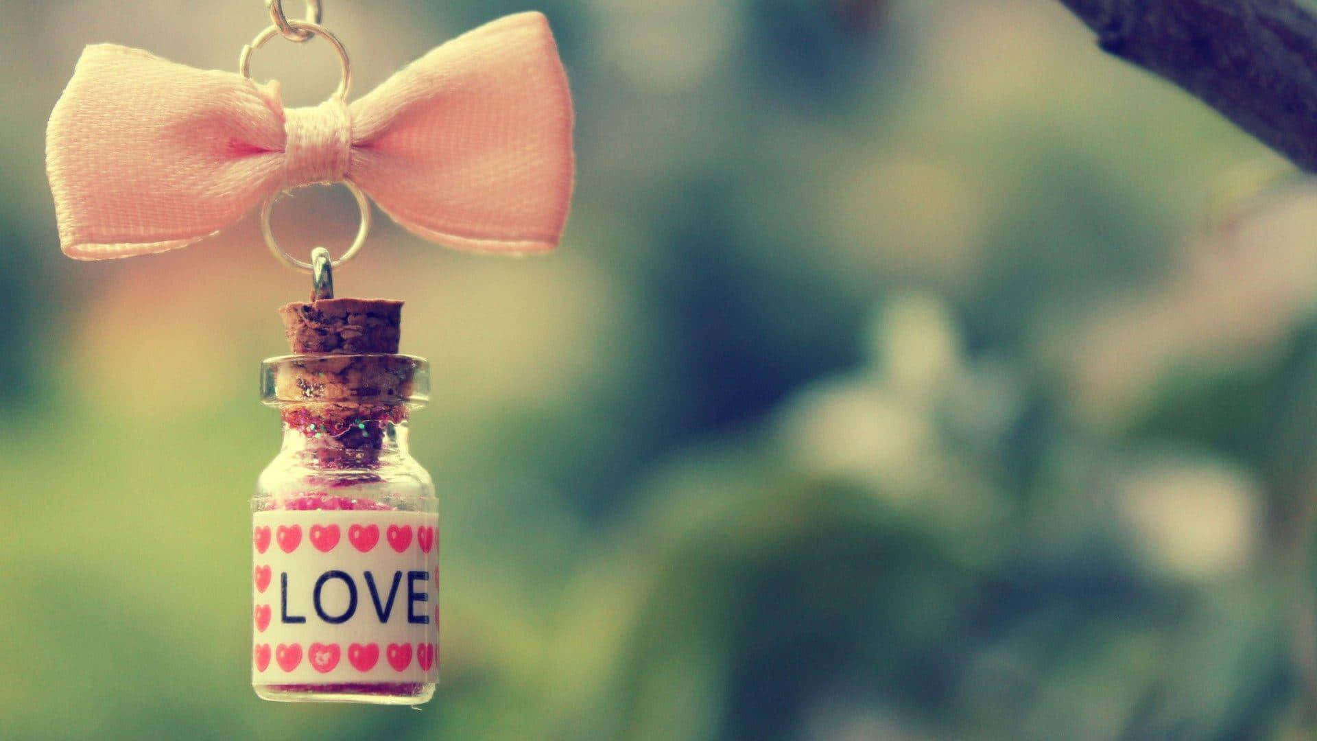 Cute Love 桌布 壁紙 — 一個精緻的粉紅色蝴蝶結裝飾著一個裝滿亮片並標有「愛」的小玻璃瓶，懸掛在柔和的模糊綠色背景前。