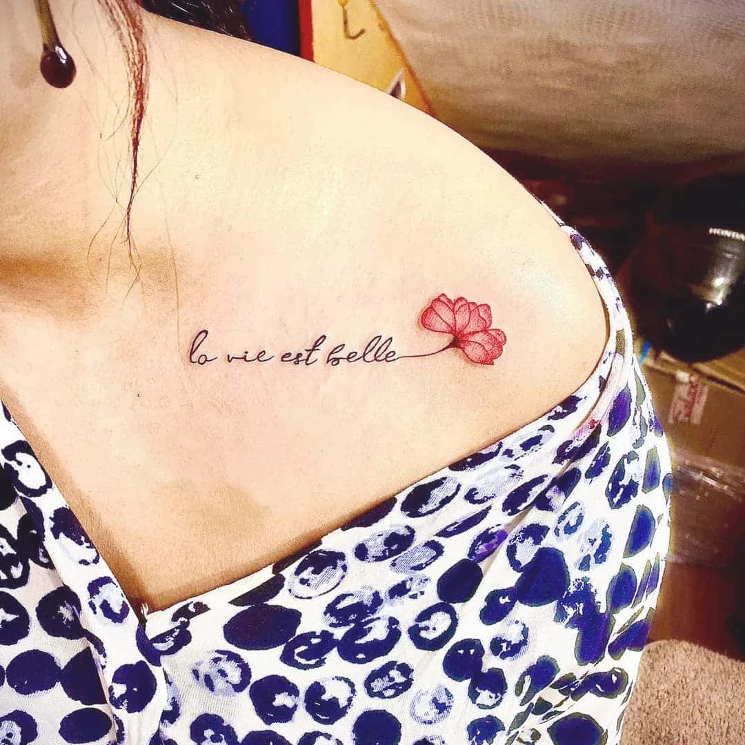 Tattoo Girl 桌布 壁紙 — 一個精緻的紅色花朵刺青，帶有草書法文文字 la vie est belle，裝飾在一個人穿著圖案上衣的肩膀上。