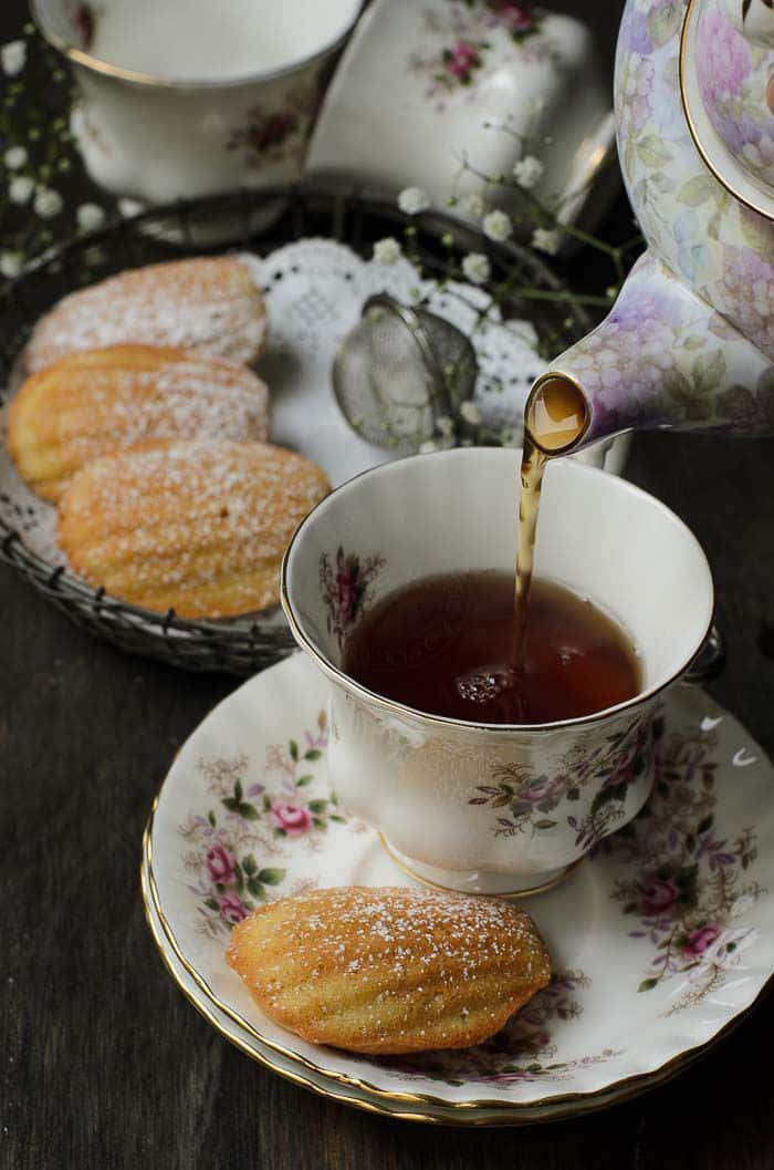 Teatime 桌布 壁紙 — 一個精緻的花卉茶壺正在將琥珀色的茶倒入一個相配的杯子中，旁邊是撒了糖粉的瑪德蓮蛋糕，放在深色木桌上。