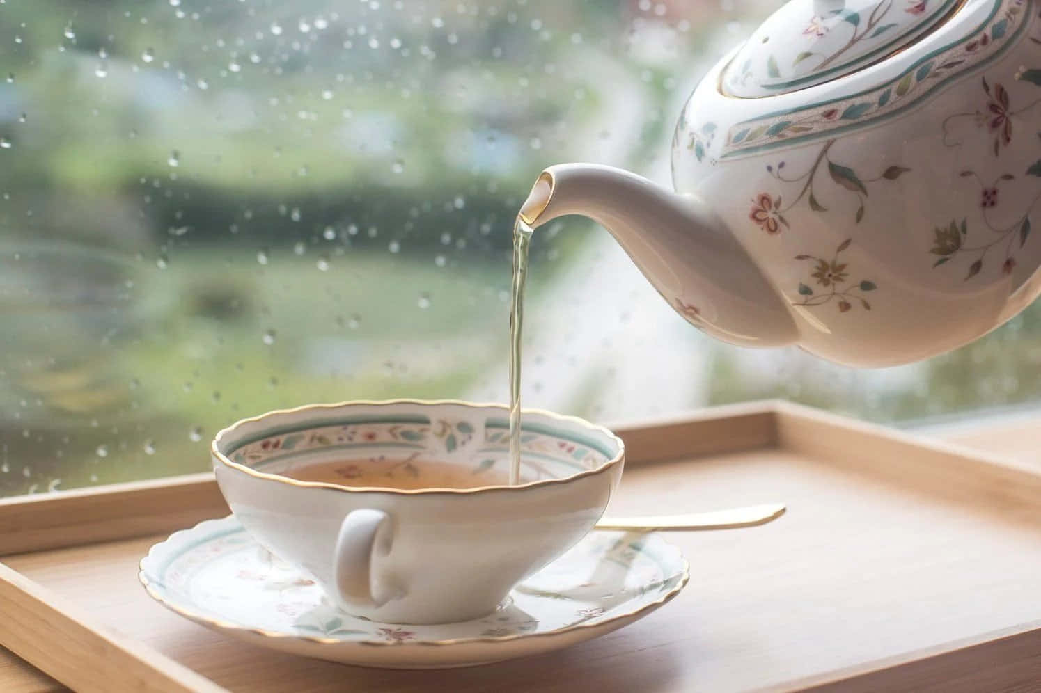 tea 桌布 壁紙 — 一個精緻的花卉茶壺正將冒煙的茶倒入一個相配的杯子中，杯子放在一個木托盤上，旁邊是佈滿雨點的窗戶。