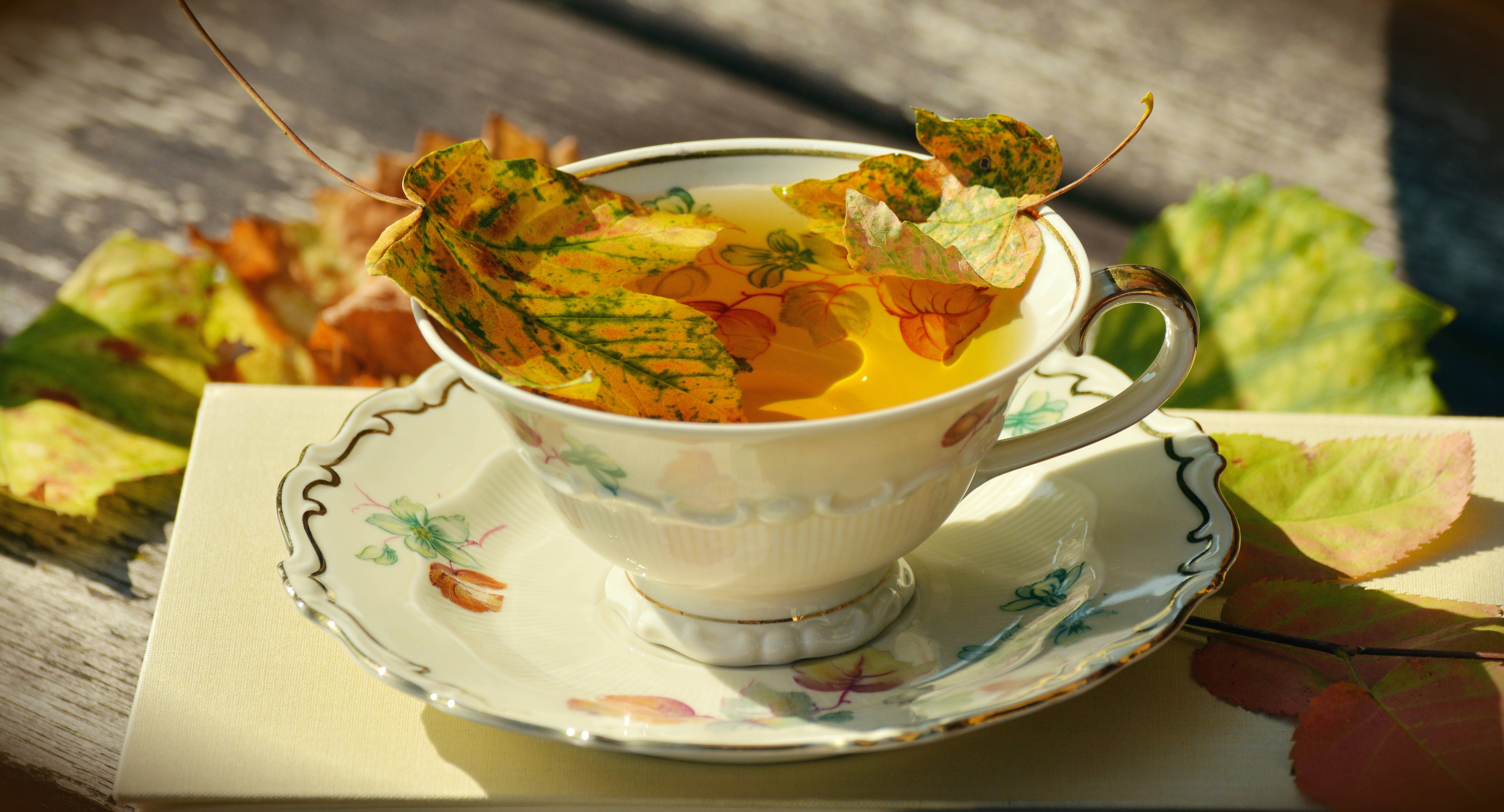 Fall Leaves 桌布 壁紙 — 一個精緻的茶杯，盛滿金黃色的茶和漂浮的秋葉，放在碟子上，旁邊是散落在木桌上的樹葉。