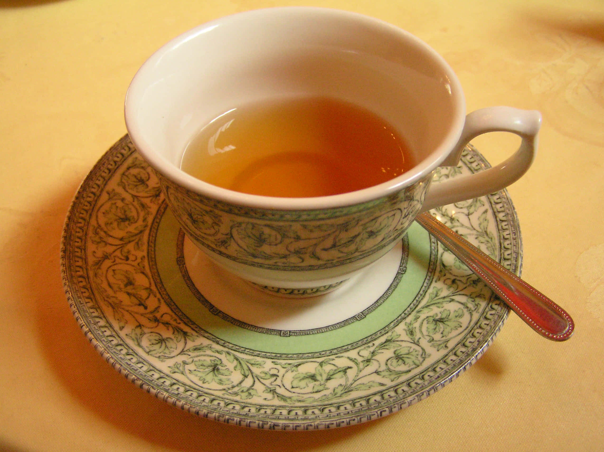 tea 桌布 壁紙 — 一個精緻的茶杯盛滿琥珀色茶，放在一個華麗的茶托上，旁邊有一把銀匙，置於金色桌布上。