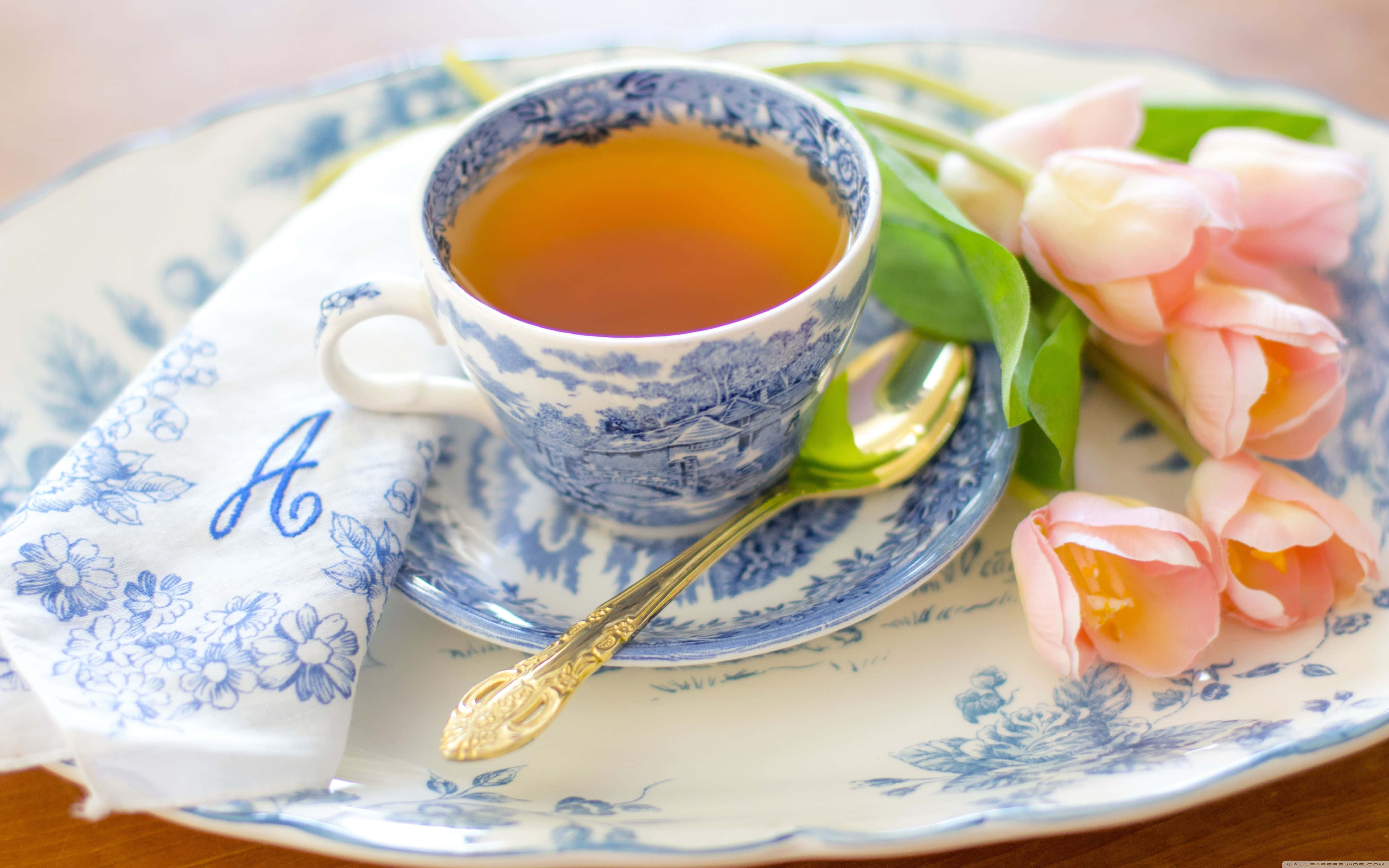 咖啡動圖 桌布 壁紙 — 一個精緻的青花瓷茶杯盛滿琥珀色茶，放在配套的茶碟上，旁邊有柔和的粉紅色鬱金香和一把金湯匙。