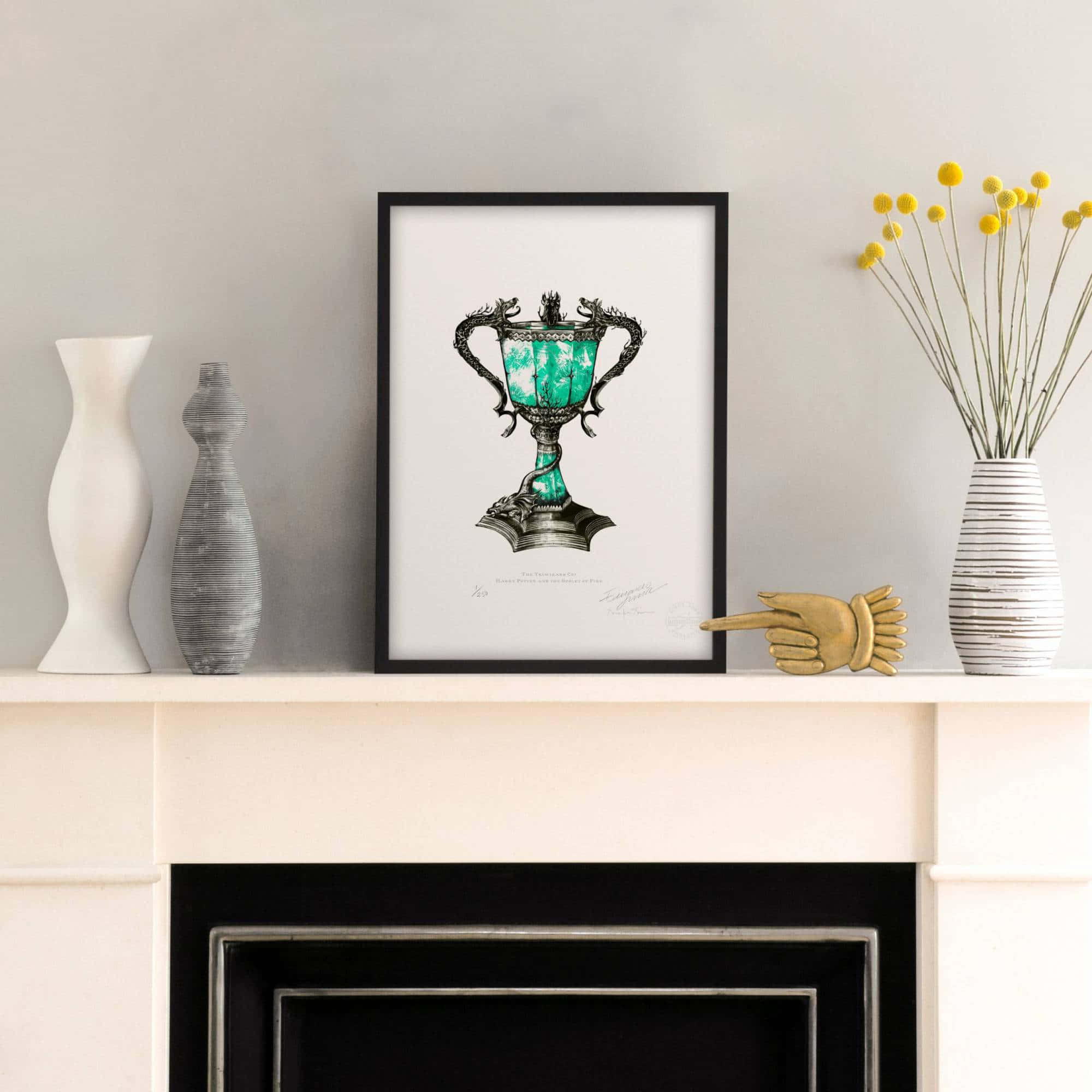 The Triwizard Cup 桌布 壁紙 — 一個精緻華麗的綠色獎盃插圖被裱在畫框中，放在白色壁爐架中央，兩側擺放著花瓶和黃色花朵。