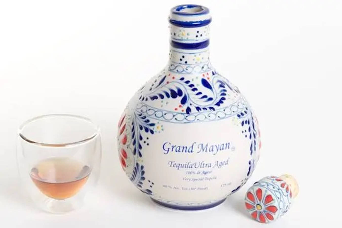 alcohol 桌布 壁紙 — 一個精美裝飾的陶瓷龍舌蘭酒瓶，帶有藍、紅、白花卉圖案，旁邊放著一杯琥珀色液體，背景為白色。