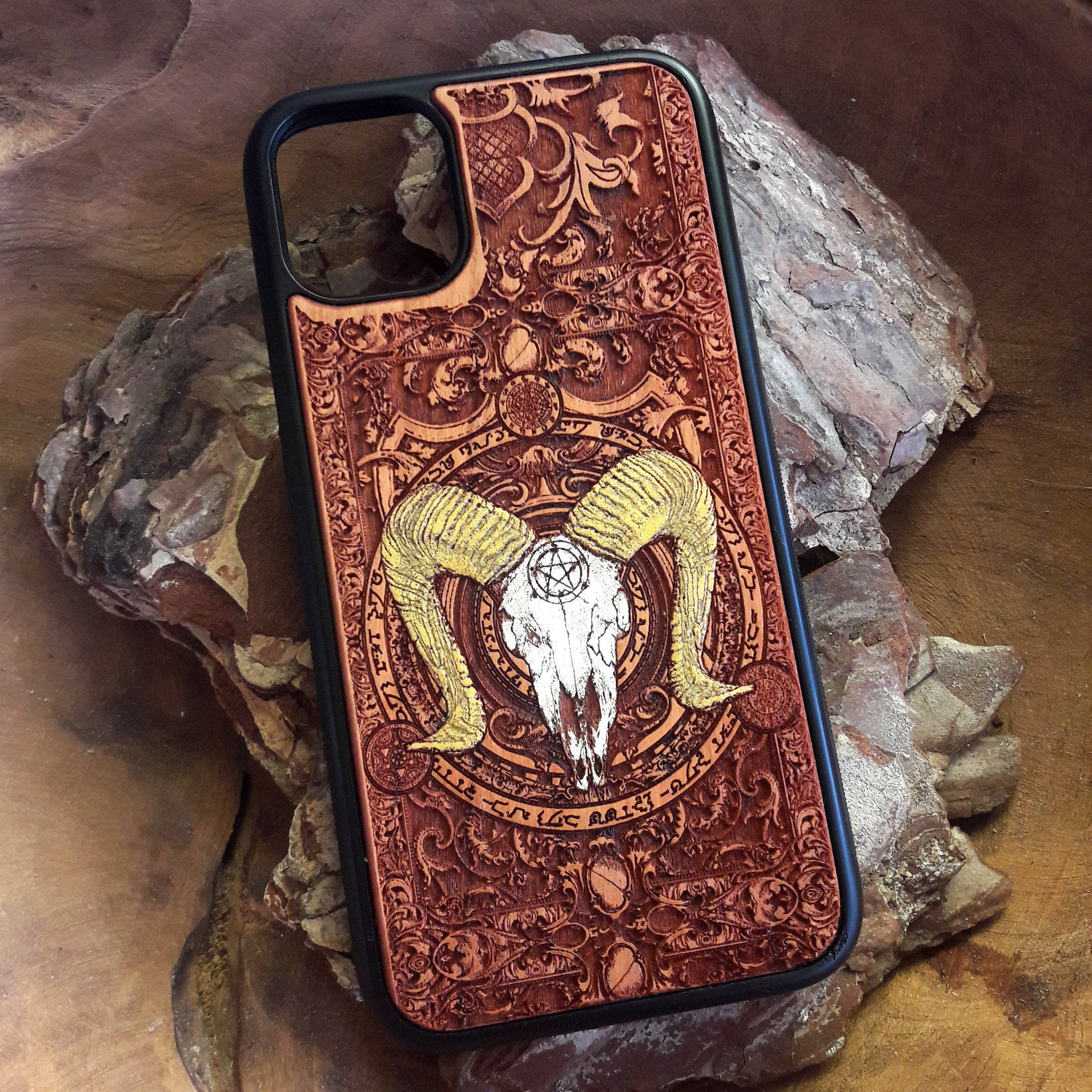 Gothic Phone 桌布 壁紙 — 一個精雕細琢的木製手機殼，帶有公羊頭骨和五角星圖案，放置在木質表面上的粗糙石頭上。
