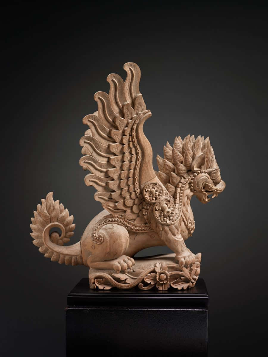 Mystical Artifacts 桌布 壁紙 — 一個精雕細琢的木製獅鷲雕像，帶有華麗的翅膀和精細的圖案，矗立在黑色底座上，背景為深色。