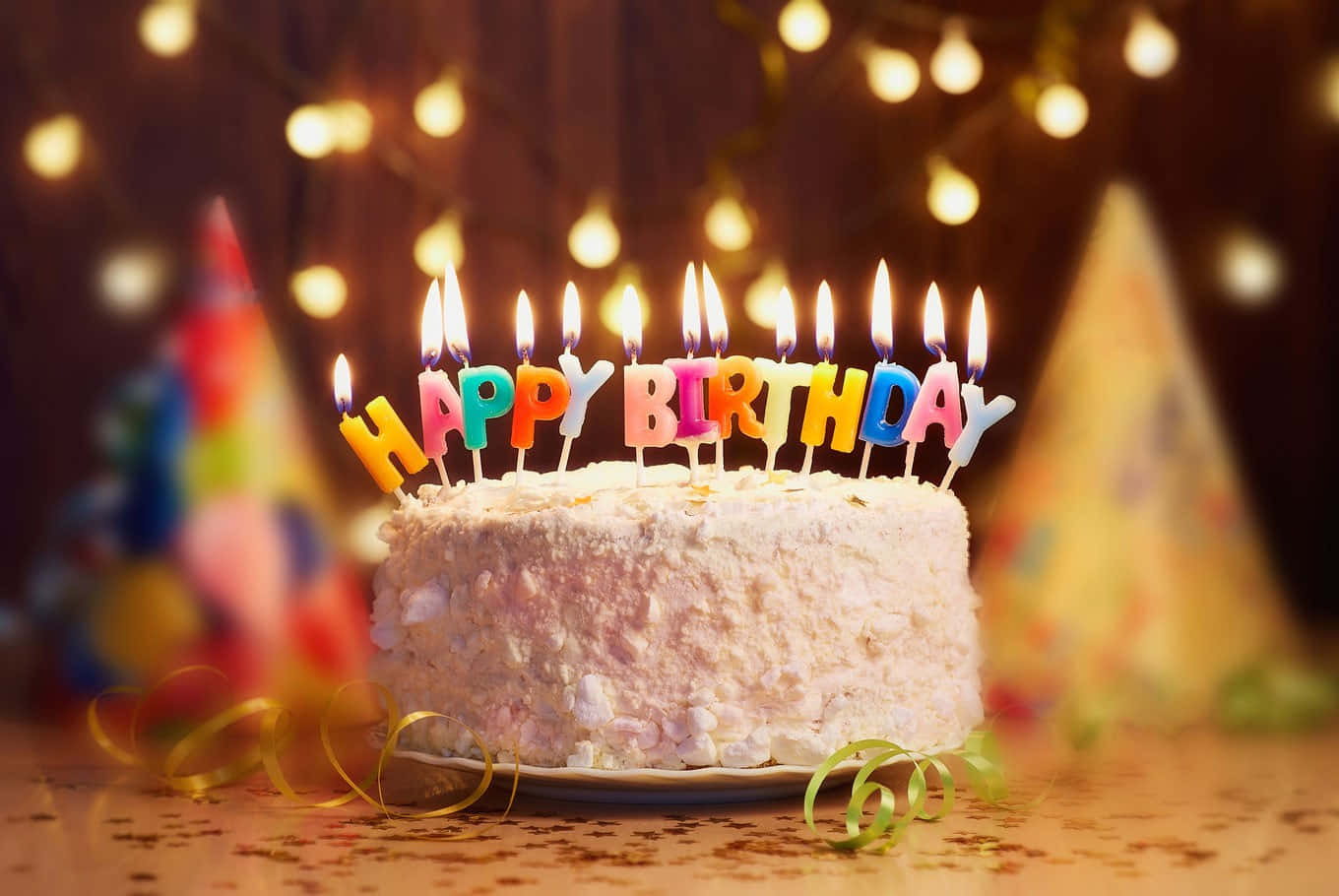 Birthday Celebration 桌布 壁紙 — 一個糖霜生日蛋糕，上面有色彩繽紛的「HAPPY BIRTHDAY」蠟燭，在溫暖的燈串和派對帽背景下閃爍。
