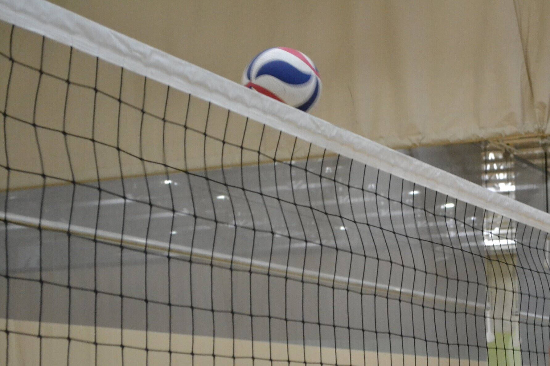 Indoor Volleyball 桌布 壁紙 — 一個紅、白、藍色的排球放在室內體育館的黑色球網頂部。
