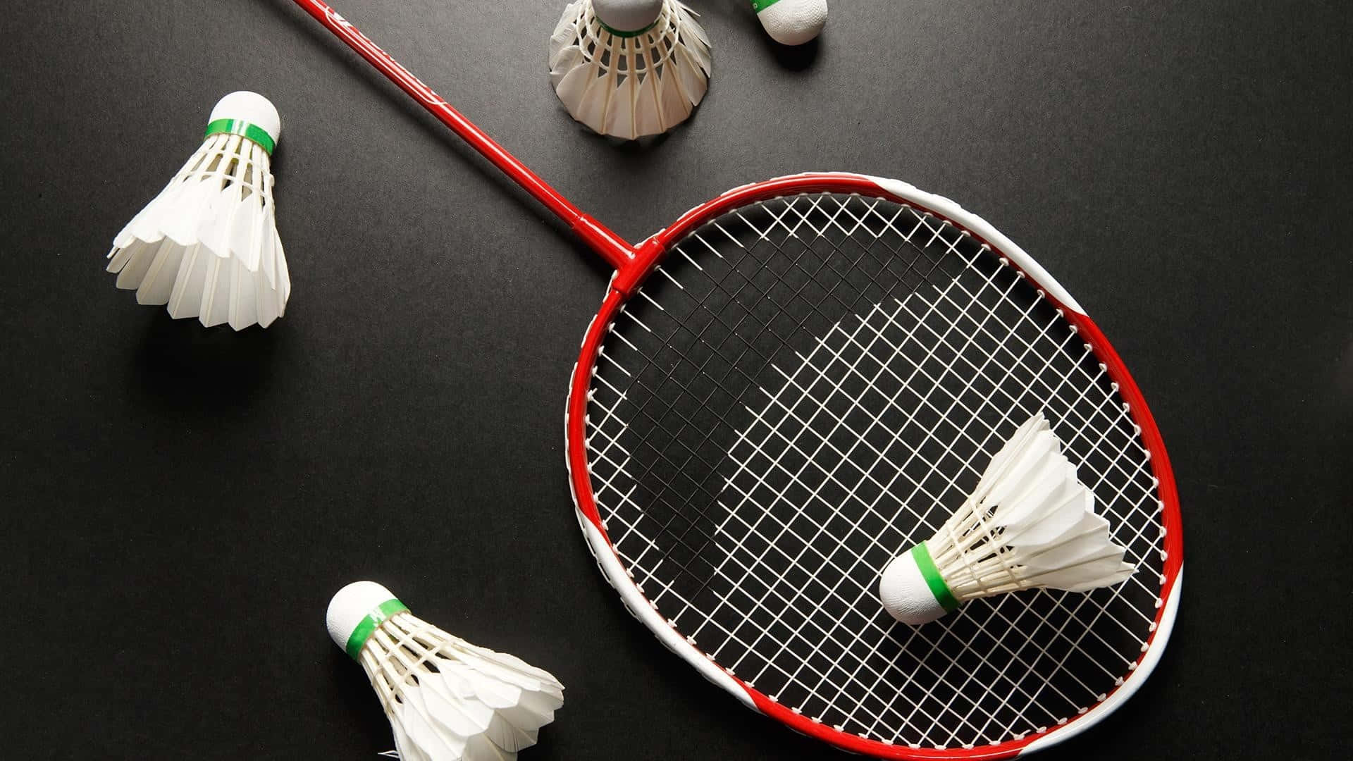 Best Badminton 桌布 壁紙 — 一個紅白相間的羽毛球拍和帶有綠色帶的白色羽毛球，排列在深色表面上。