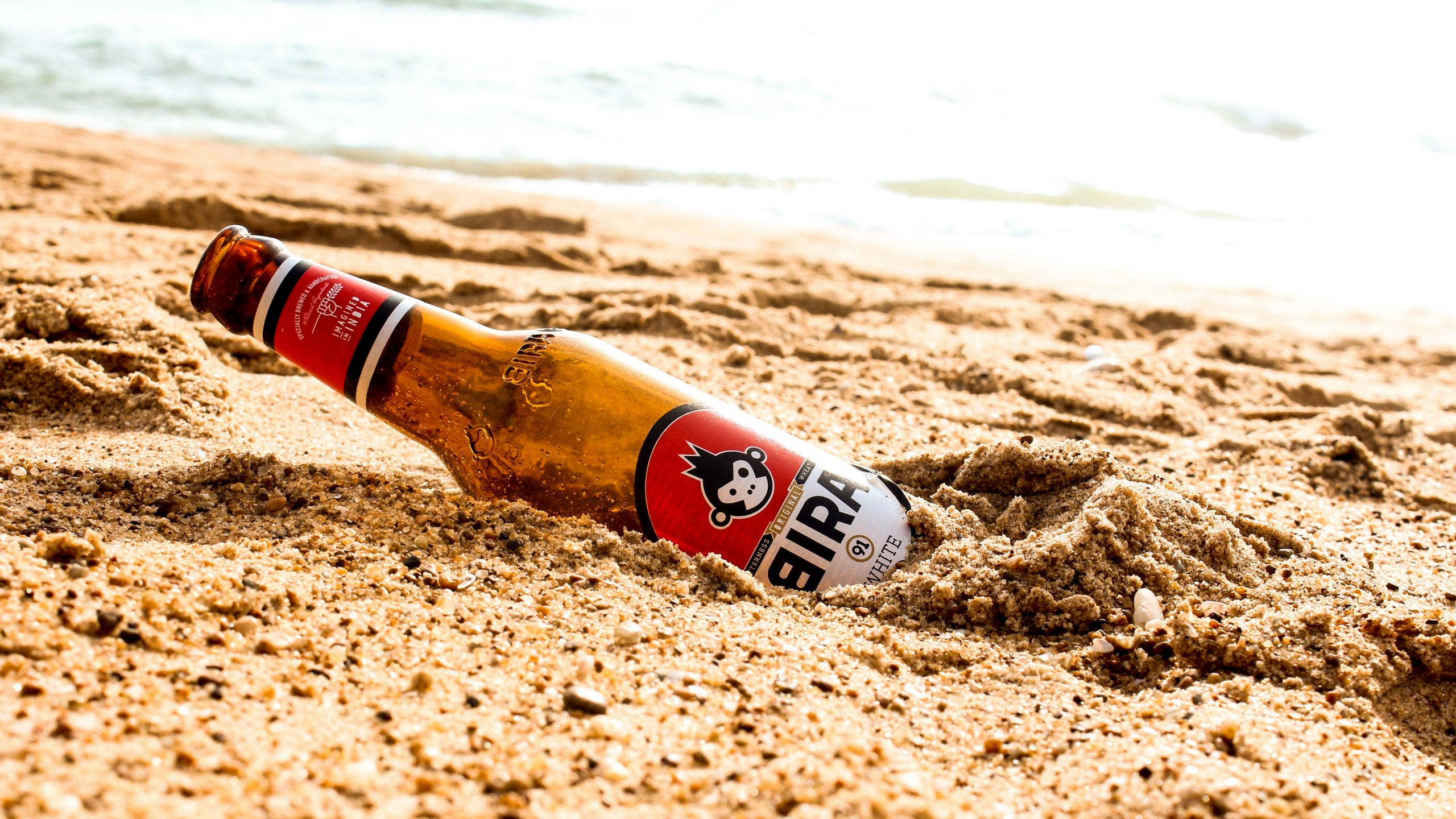 Beer 桌布 壁紙 — 一個紅白色的Bira啤酒瓶半埋在近海邊的金色沙中。