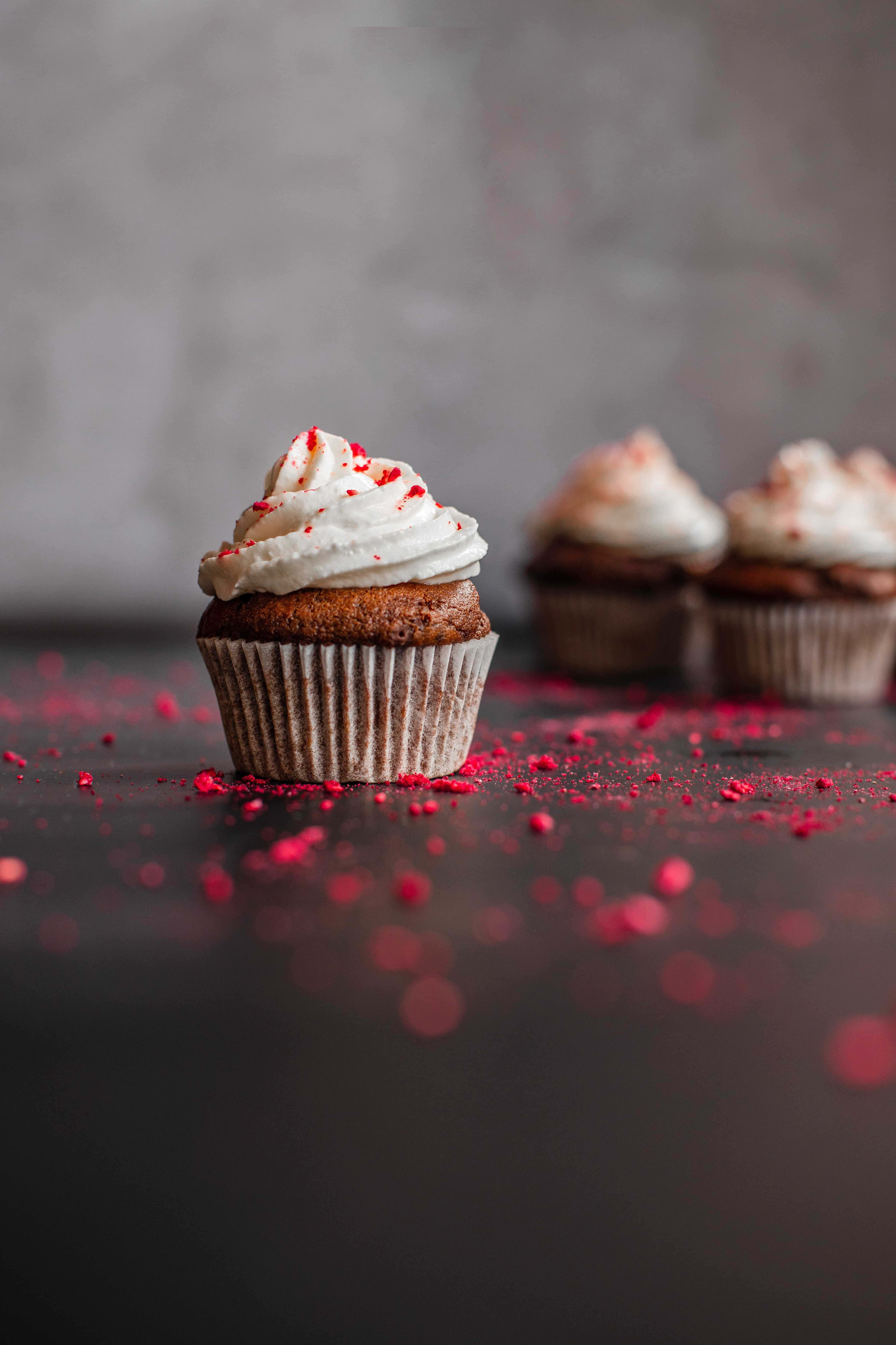 Cupcake 桌布 壁紙 — 一個紅絲絨杯子蛋糕，上面有白色糖霜和紅色糖粒，聚焦在深色表面上，背景的杯子蛋糕模糊不清。