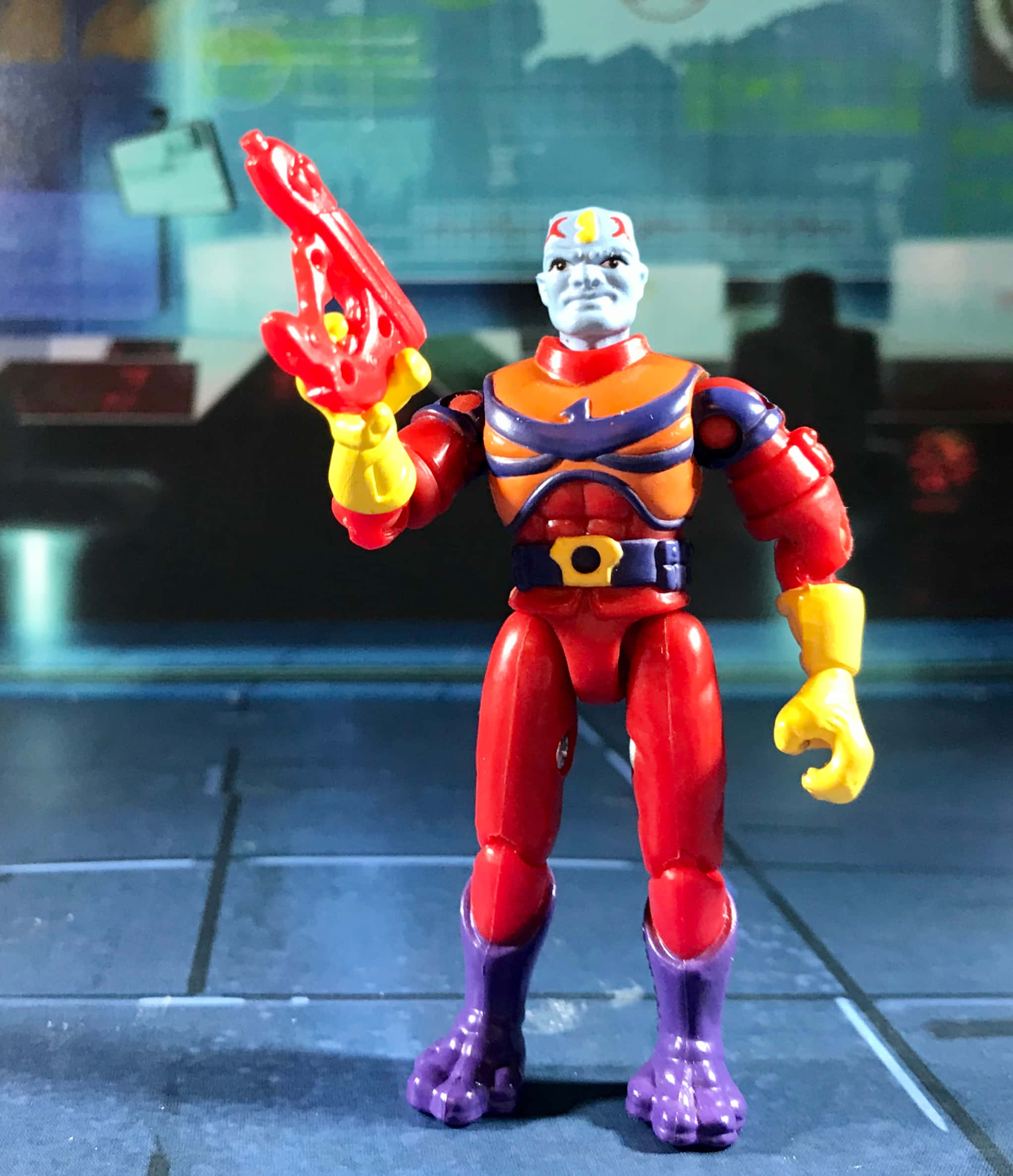 toys 桌布 壁紙 — 一個紅色、橙色和紫色的動作人偶，手持黃色武器，站在瓷磚地板上，背景是模糊的未來風格。
