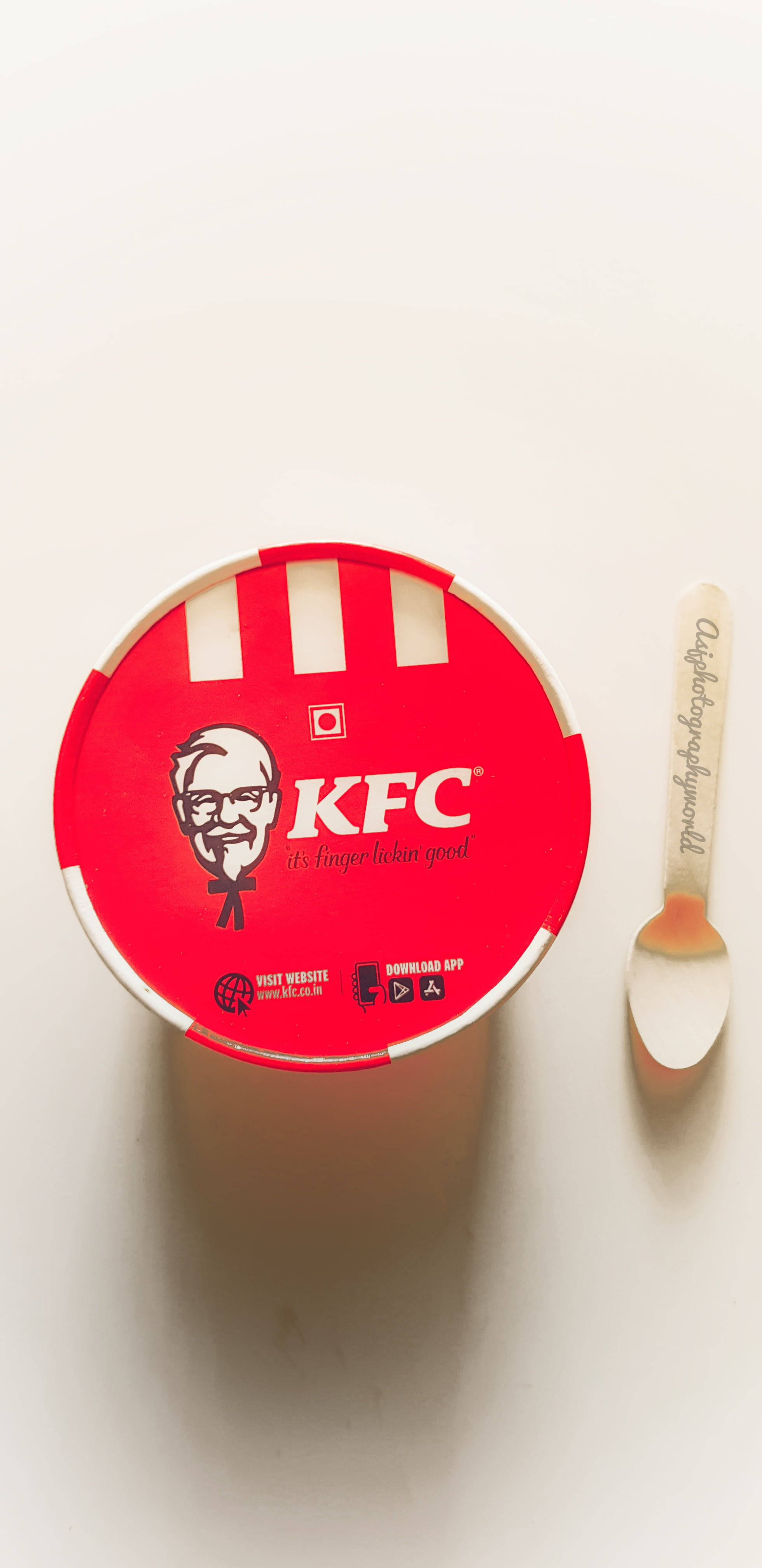 Kfc 桌布 壁紙 — 一個紅色KFC桶，上面有桑德斯上校標誌和品牌湯匙，放在純白色表面上，從正上方俯瞰的視角。