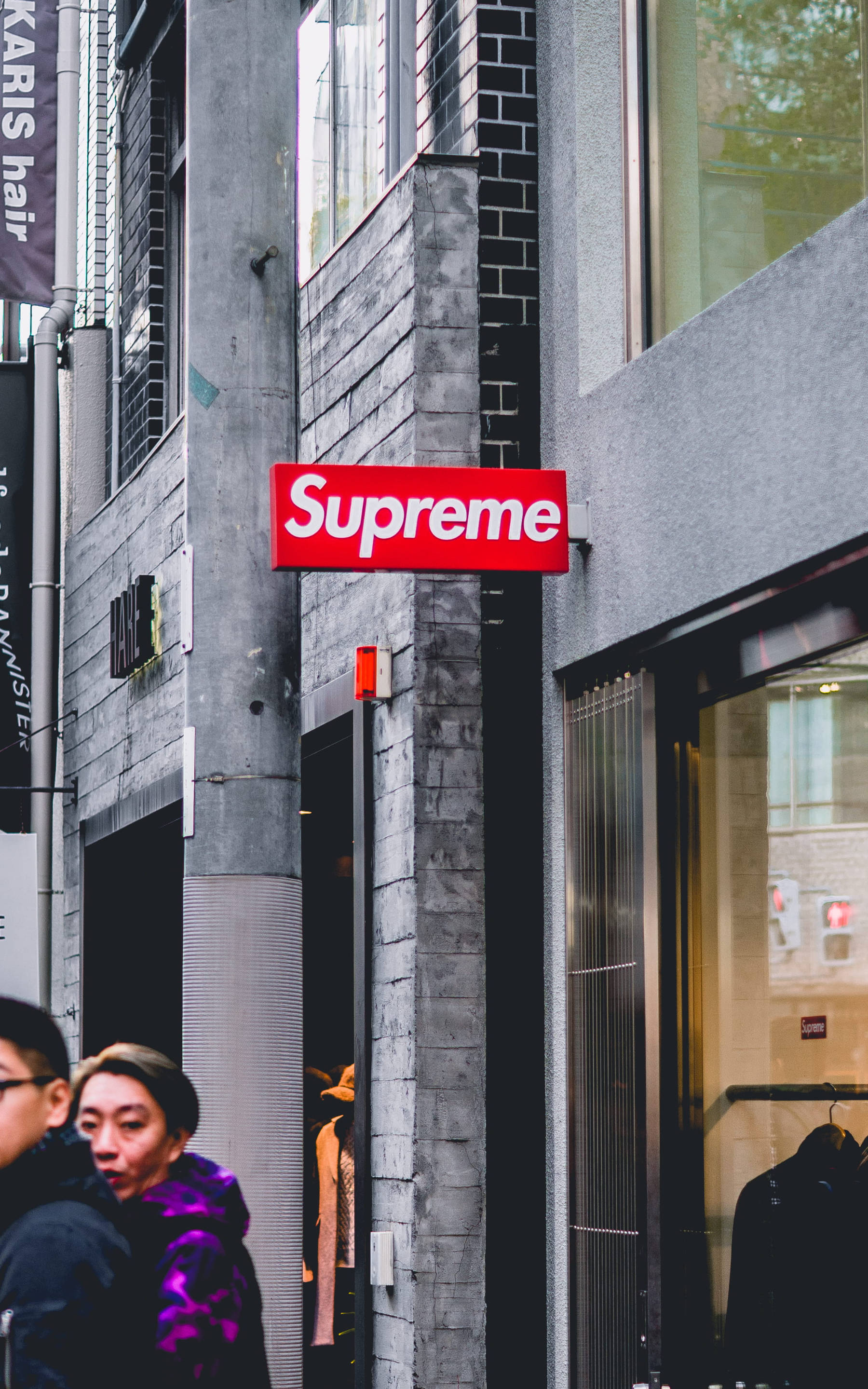 Dope Supreme 桌布 壁紙 — 一個紅色Supreme標誌掛在灰色磚建築上，位於繁忙的城市街道，有行人經過。