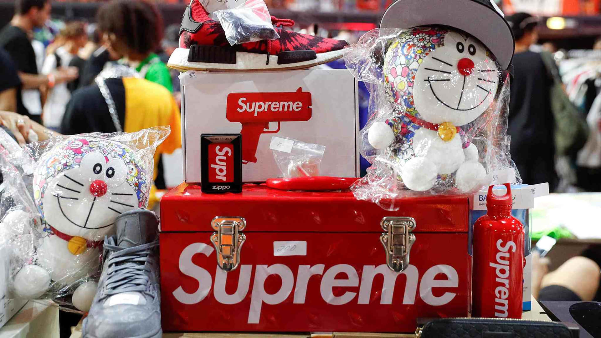 supreme 桌布 壁紙 — 一個紅色Supreme行李箱展示了一系列品牌商品，包括哆啦A夢毛絨玩具、運動鞋和Zippo打火機，在一個擁擠的快閃店內。