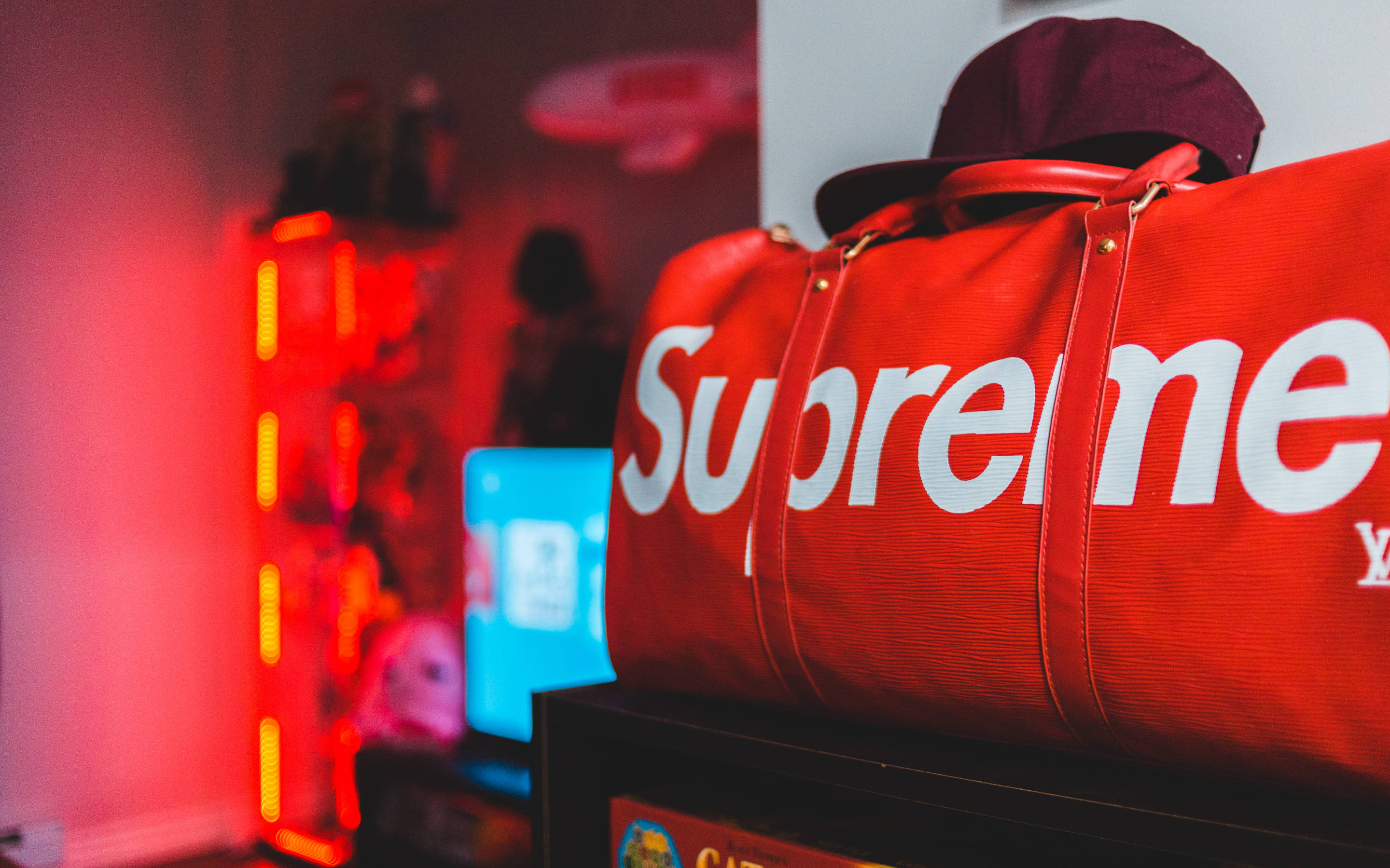 超屌 Supreme 桌布 壁紙 — 一個紅色Supreme行李袋和一個栗色帽子放在一個架子上，沐浴在來自發光垂直條紋的溫暖紅色霓虹燈光中。